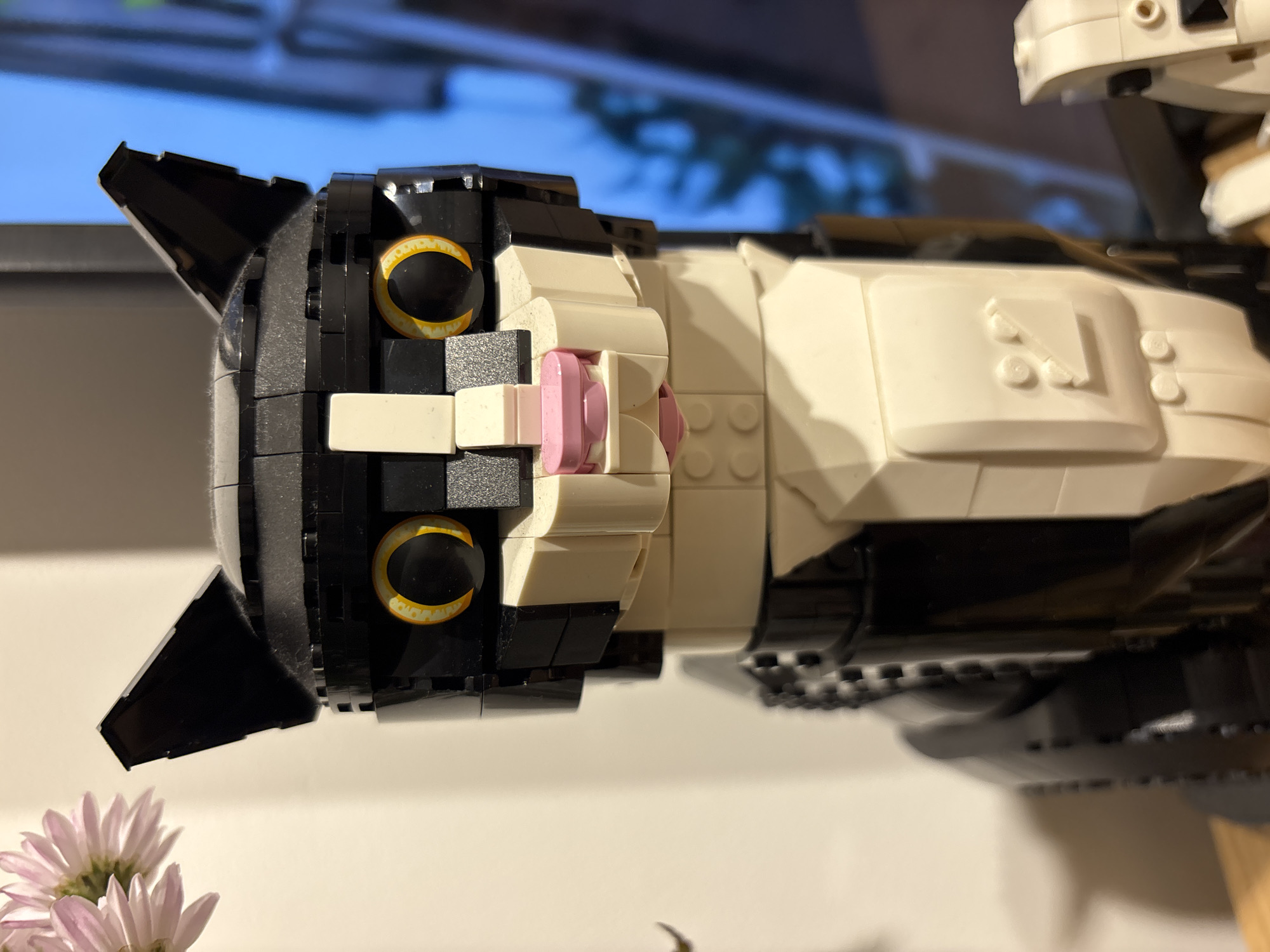 A close-up shot depicts a Lego model of a tuxedo cat. The cat has black and white coloration. Its eyes are black with golden yellow irises. The nose is pink. The cat is resting against a white background. In the foreground, there are some pink chrysanthemums.