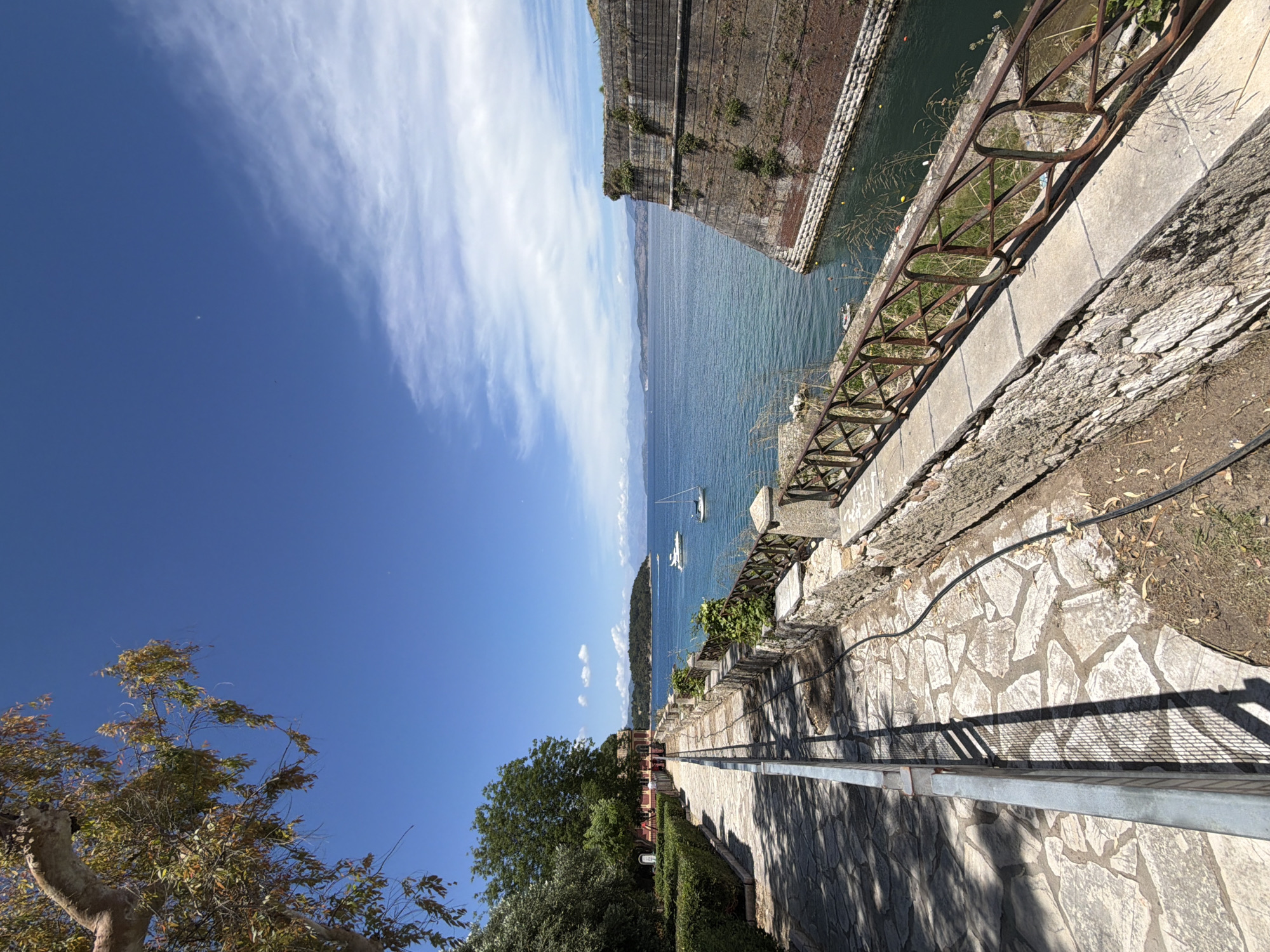 This vertically-oriented image, set in Corfu, Greece, captures a scenic coastal view under a bright, clear sky. A vast expanse of calm, deep blue sea occupies the left side of the frame, with two white sailboats visible on its surface. Along the upper left, a formidable, ancient-looking stone fortification wall, covered in patches of greenery, rises directly from the water, showcasing historical architecture.
On the right, a winding pathway paved with irregular stone slabs extends into the distance. This path is bordered by a metal railing, which transitions from a rusty, older style in the foreground to a newer, silver appearance further along. Prominent shadows from the railing and overhead foliage are cast sharply across the sunlit path, indicating a clear, sunny day, likely mid-morning or afternoon. Lush green trees and hedges line parts of the pathway, adding natural elements to the scene. In the far distance along the path, small, indistinct human figures can be seen, suggesting people are walking and enjoying the view.
The sky above is a vibrant blue, featuring a band of wispy white clouds. A faint, distant mountainous landmass stretches across the horizon, adding depth to the panorama. The scene conveys a sense of peaceful historical beauty. No visible text is present in the image.
