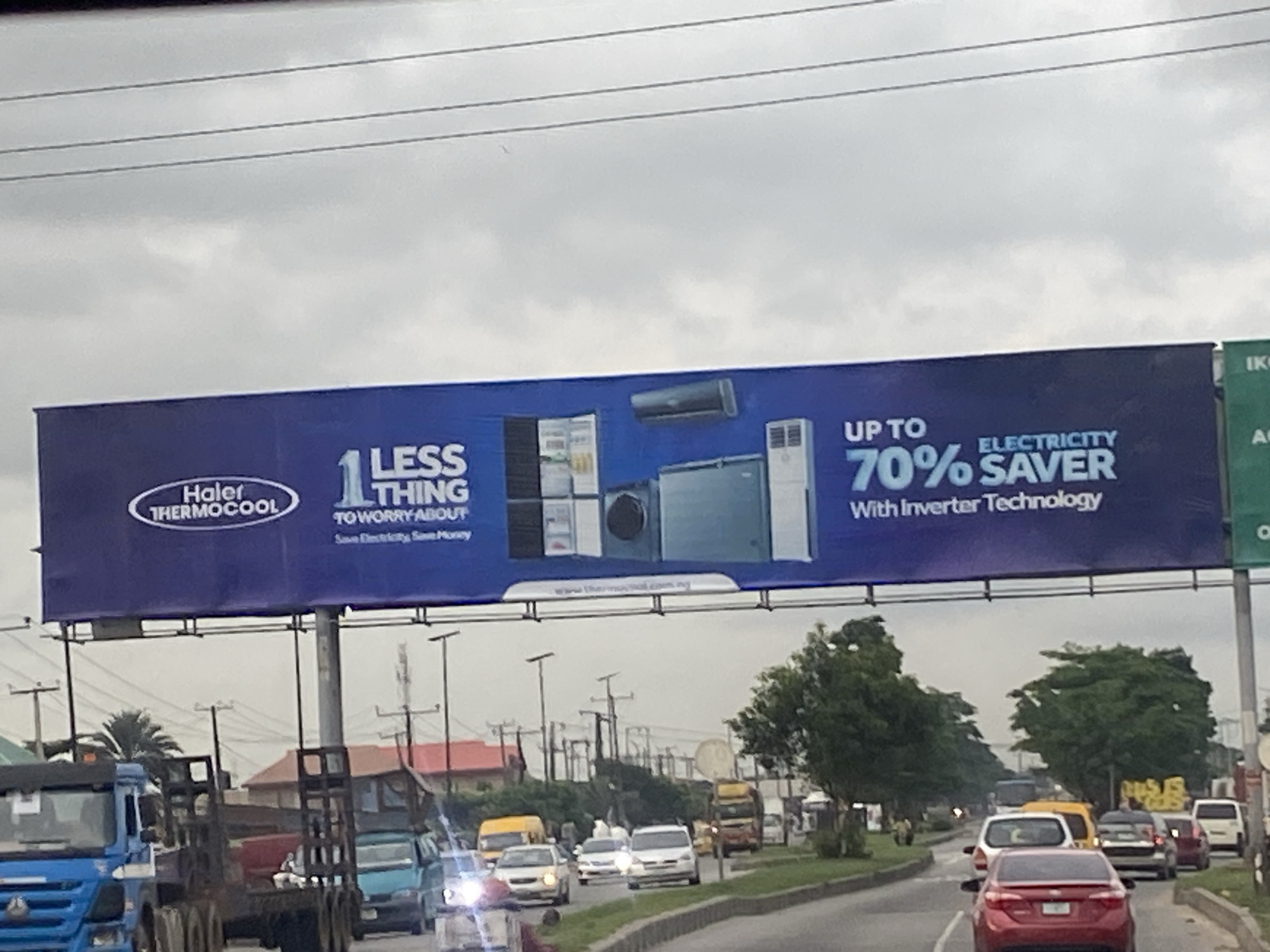 A wide-angle, eye-level, slightly blurry photograph shows a billboard advertising Haier Thermocool appliances in Ikeja, Nigeria. The billboard is positioned above a busy road filled with various vehicles including trucks, cars, and buses. The sky is overcast and grey.
The billboard is predominantly blue and features an image of several appliances, including what appears to be a refrigerator, an air conditioning unit, and a washing machine. Text on the billboard reads "Haier THERMOCOOL" in an oval logo. Below this, it says "1 LESS THING TO WORRY ABOUT" and "Save Electricity, Save Money." To the right, it advertises "UP TO 70% ELECTRICITY SAVER With Inverter Technology." A website address, "www.thermocool.com.ng," is visible at the bottom of the billboard.
On the road, a blue truck with a flatbed is in the foreground on the left. Other vehicles, including a red sedan, white vans, and yellow buses, are visible in traffic. Utility poles and power lines are present along the roadside, as are trees and some buildings. The scene appears to be captured from another vehicle moving through the area.