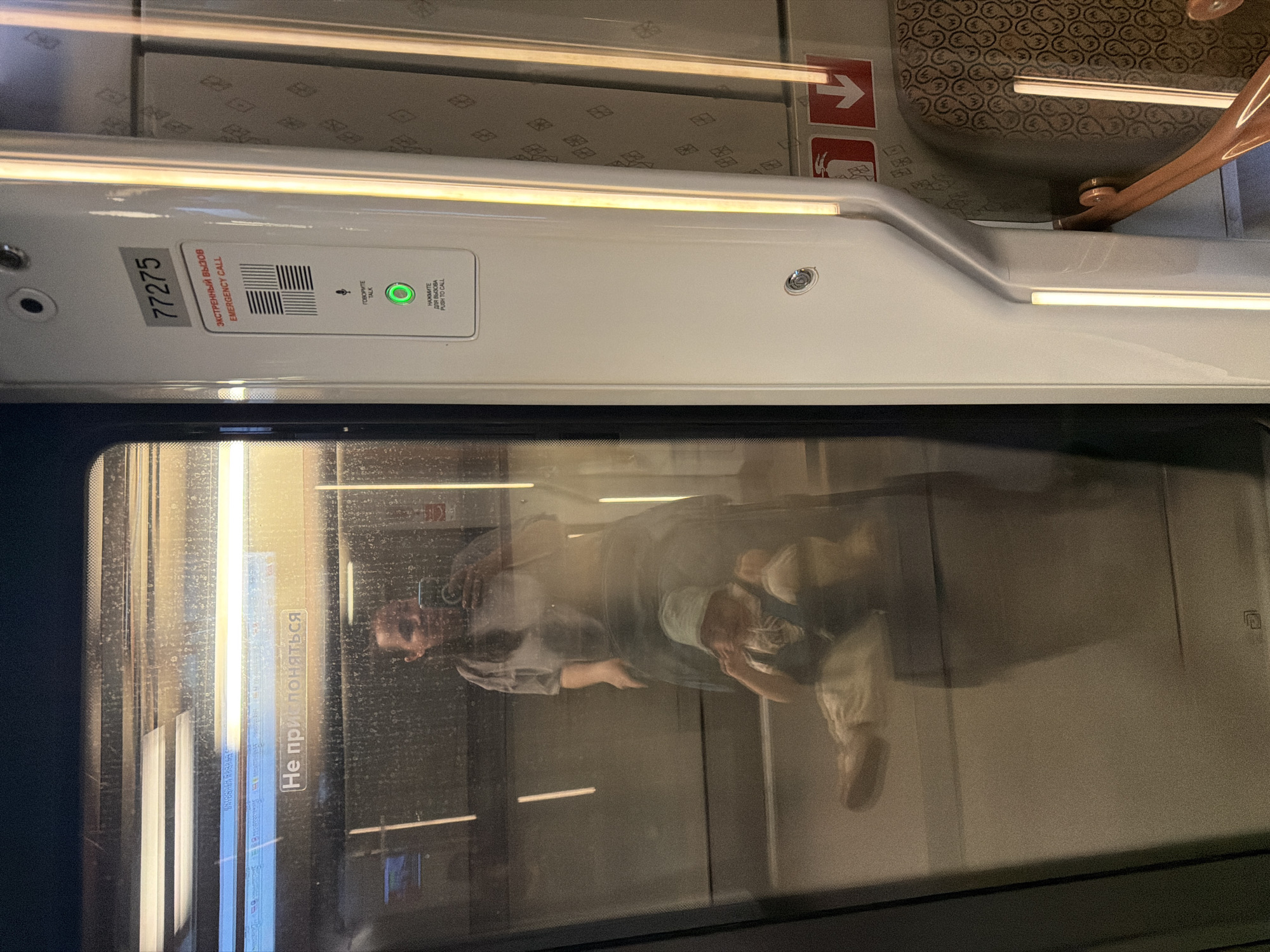 The image shows the interior of a train carriage, possibly within the Moscow, Russia, metro system, as evidenced by Russian text on stickers and signs. The carriage is mostly white, and visible is an emergency call button panel labeled in both Russian and English. Reflections in the window show a person holding a smartphone and a possible child, both in lighter-colored clothing. The window also features a warning sticker with Russian text that reads "Не прислоняться."