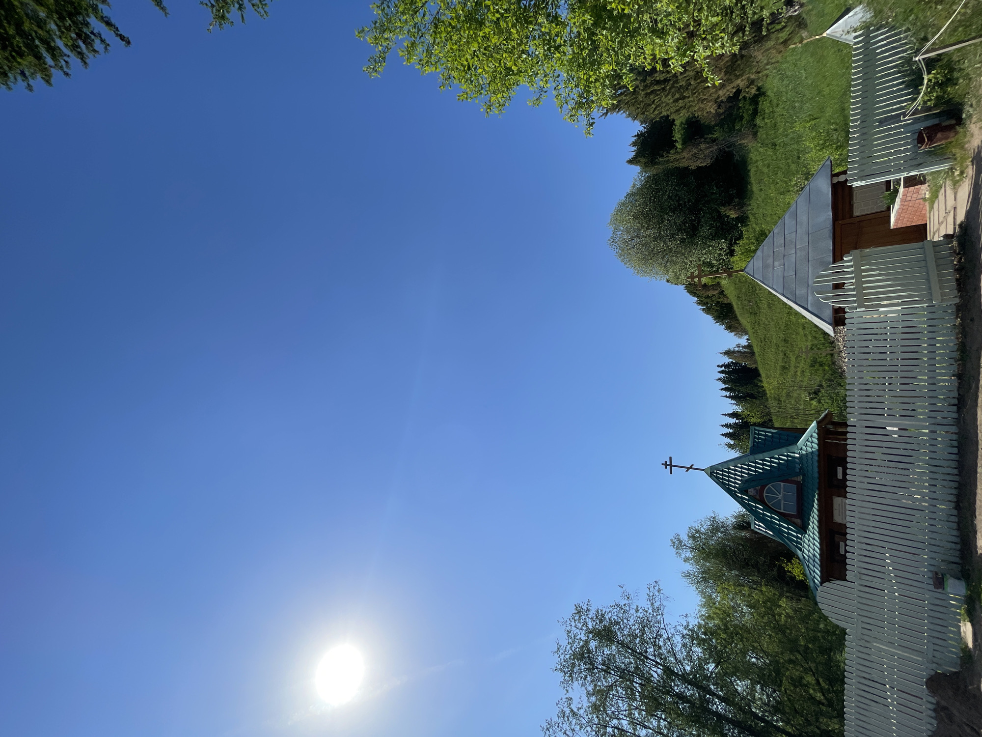 The image shows a building with a turquoise roof and a cross at the top, located in Перевозное, Russia. The building is surrounded by a white fence. The sky is blue and clear with the sun shining brightly. Trees and grass are visible in the background.