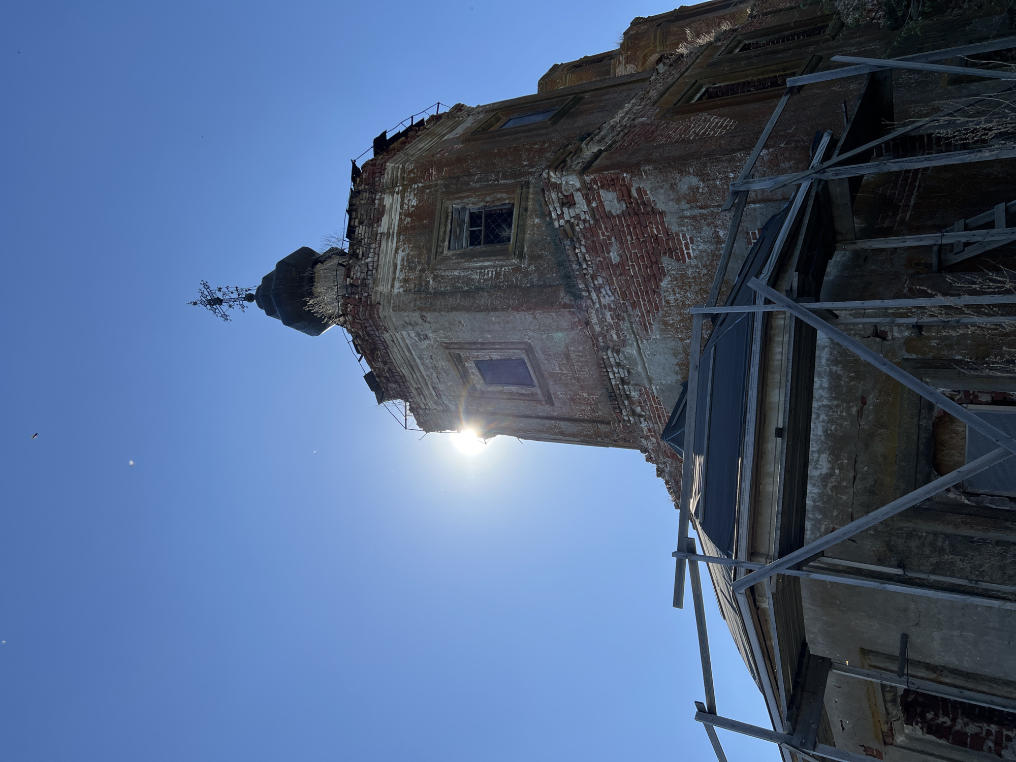 The image shows a low-angle view of a partially ruined brick building in Шуран, Russia, set against a clear blue sky. The building's facade is weathered, with significant deterioration of the brickwork and plaster. Scaffolding supports sections of the building. The sun shines from behind the structure. A dome with a cross sits atop a square tower.