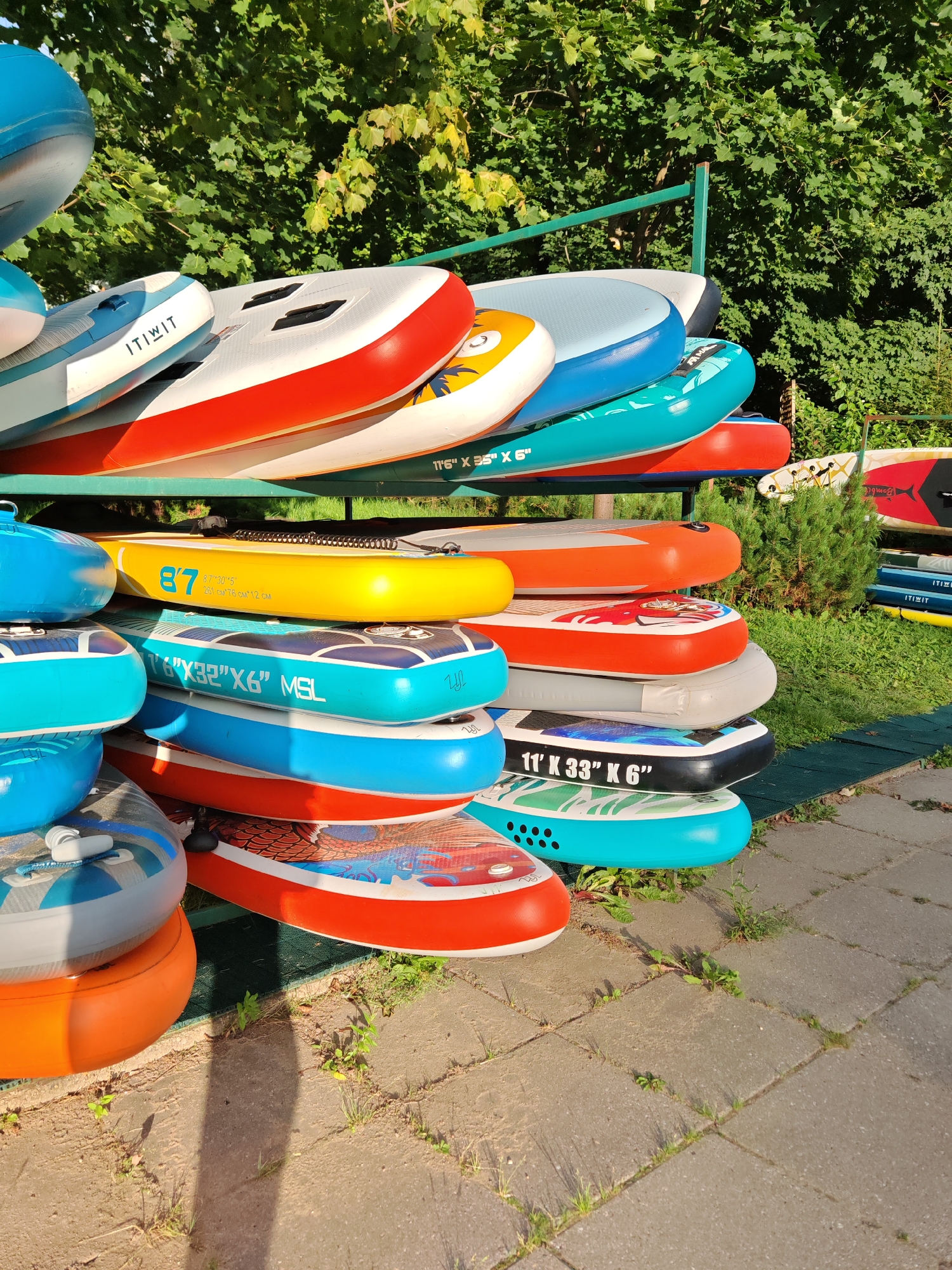 In Rosinka, Russia, a row of inflatable stand-up paddleboards (SUPs) are neatly stacked on a metal rack outdoors, under the shade of lush green trees. The paddleboards vary in color and design, with some featuring bright oranges, blues, yellows, and reds, while others have more subtle patterns. Some boards display branding and dimensions such as "ITIWIT," "11'6" X 35" X 6"," and "8'7" 30.5" X 6" 25 CM 76 CM X 12 CM". The SUPs are arranged in multiple tiers, creating a colorful display. In the background, more paddleboards are visible, also stacked on racks. The ground in front of the racks is paved with interlocking stones and sparse vegetation. The scene appears to be a rental or storage area for water sports equipment, possibly near a body of water, given the context of Rosinka's location. There are no people visible in the image.