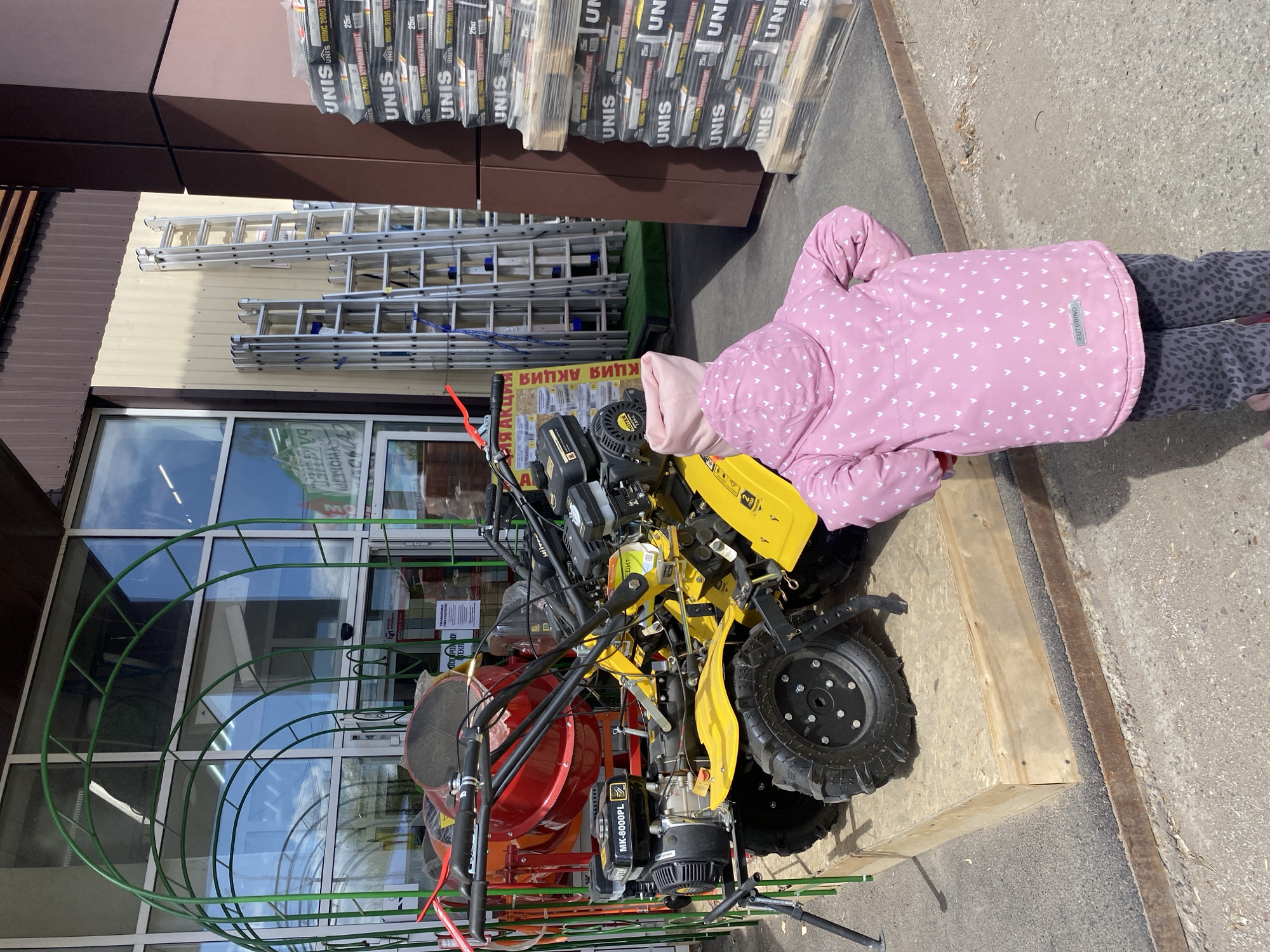 Here's a detailed description of the image:
**Scene:** The image appears to be taken outdoors in front of a building that is likely a hardware or construction supply store in the city of Chaikovsky, Russia.
**Subjects and Objects:**
* A young child wearing a pink jacket with white hearts is looking at some equipment.
* The equipment includes a bright yellow tiller/cultivator and a red concrete mixer, both of which appear to be new and for sale.
* Ladders, stacks of bagged construction materials labeled "UNIS" in Cyrillic, and a green arched frame (possibly for a greenhouse) are visible behind the equipment.
* The building behind has large windows.
**Environment:**
* The area is paved.
* The store has brown and beige facade paneling.
**Actions and Interactions:**
* The child is closely observing the tiller/cultivator.
* The equipment is displayed outside the store, likely for sale.