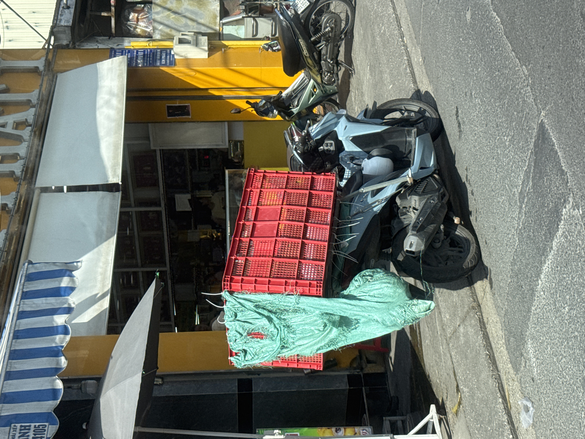 A light blue motorcycle is parked on the side of a street in Thủ Đức, Vietnam. The motorcycle is carrying a large red plastic crate strapped to its rear. A green tarp is draped over the crate and extends downwards, appearing to be tied to the motorcycle. The crate is full of items, though their contents are not clearly visible. Behind the motorcycle, a darker green and black motorcycle is also parked. The street has a grey concrete sidewalk and an asphalt road. Buildings with yellow facades and blue and white striped awnings are visible in the background. The scene appears to be a commercial area with shops or storefronts.