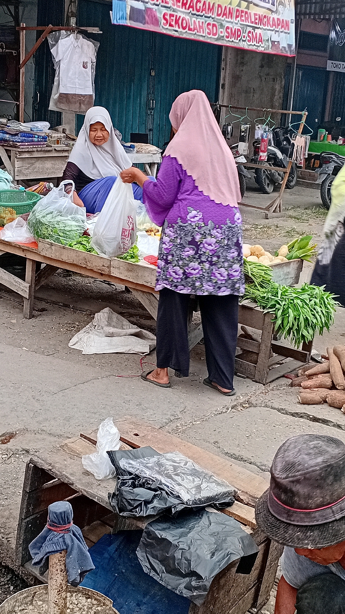 This image depicts an outdoor market scene in Jambi, Indonesia. In the foreground, two women are interacting at a stall. One woman, wearing a white hijab and a dark top, is seated and holding a plastic bag. The other woman, wearing a pink hijab and a purple floral-patterned top, is standing and appears to be assisting the seated woman. They are surrounded by various goods, including fresh produce like leafy green vegetables and what appear to be potatoes and yams. The stall is set up on wooden planks, with other market stalls and motorcycles visible in the background. A banner with Indonesian text, likely advertising school uniforms, is also present. The ground is paved with concrete. A second person, wearing a hat, is partially visible in the lower right corner of the image. The overall impression is of a bustling local market on a cloudy day.