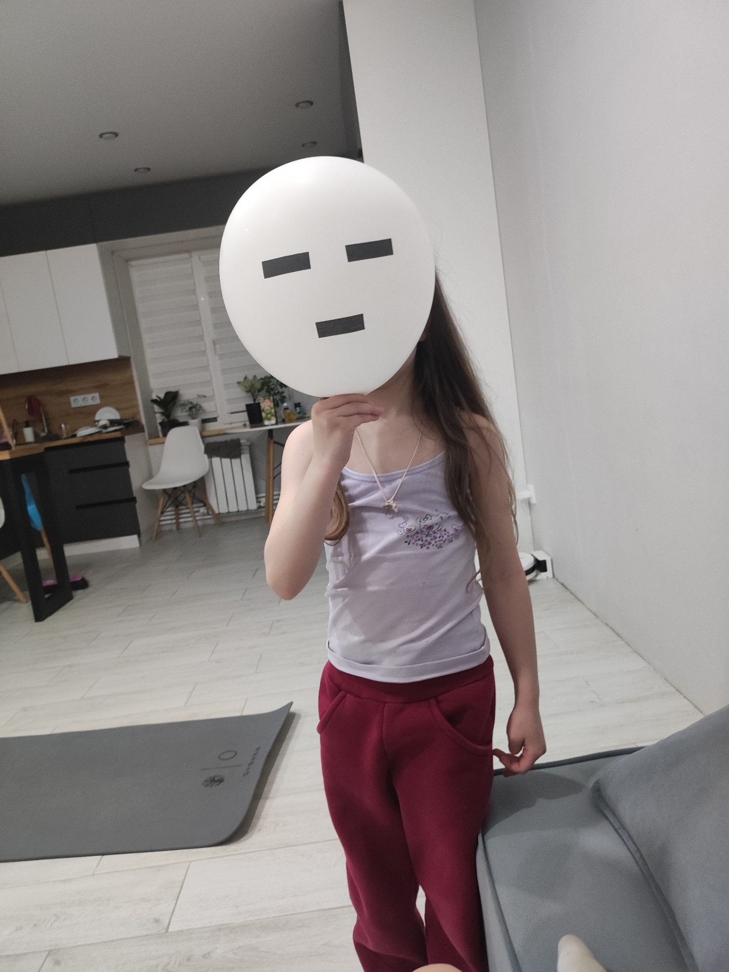 A young girl is standing indoors, holding a large white balloon with a simple, blocky face drawn on it, obscuring her own face. She is wearing a light purple tank top with a floral design and maroon sweatpants. She wears a necklace with a small pendant. The interior appears to be a modern apartment. In the background to the left, there is a kitchen area with light-colored cabinets and dark gray lower cabinets. A white chair sits near a table with plants on it. A gray yoga mat is on the floor in the foreground. Part of a gray sofa is visible on the right. There is no discernible information in the image to place the location within the city of Votkinsk, Russia.