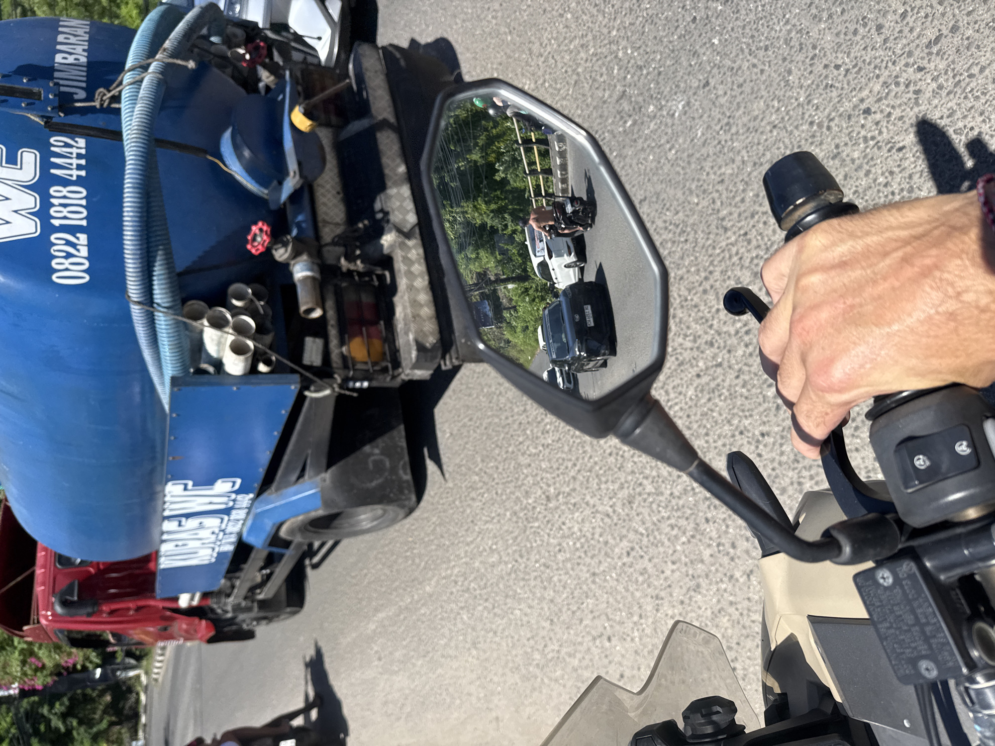 From the perspective of a motorcycle rider on an asphalt road in Kuta Selatan, Indonesia, a blue water truck, with the company name "WC Jimbara" and phone number "0822 1818 4442" printed on its side, is seen in the left side of the frame. The motorcycle's right handlebar, complete with grip and brake lever, is visible in the lower right corner, held by a right hand. Reflected in the motorcycle's mirror is a view of the road behind, showing a motorcyclist following a white car and a black van. Trees line the side of the road in the mirror's reflection. The street appears to be in good condition.