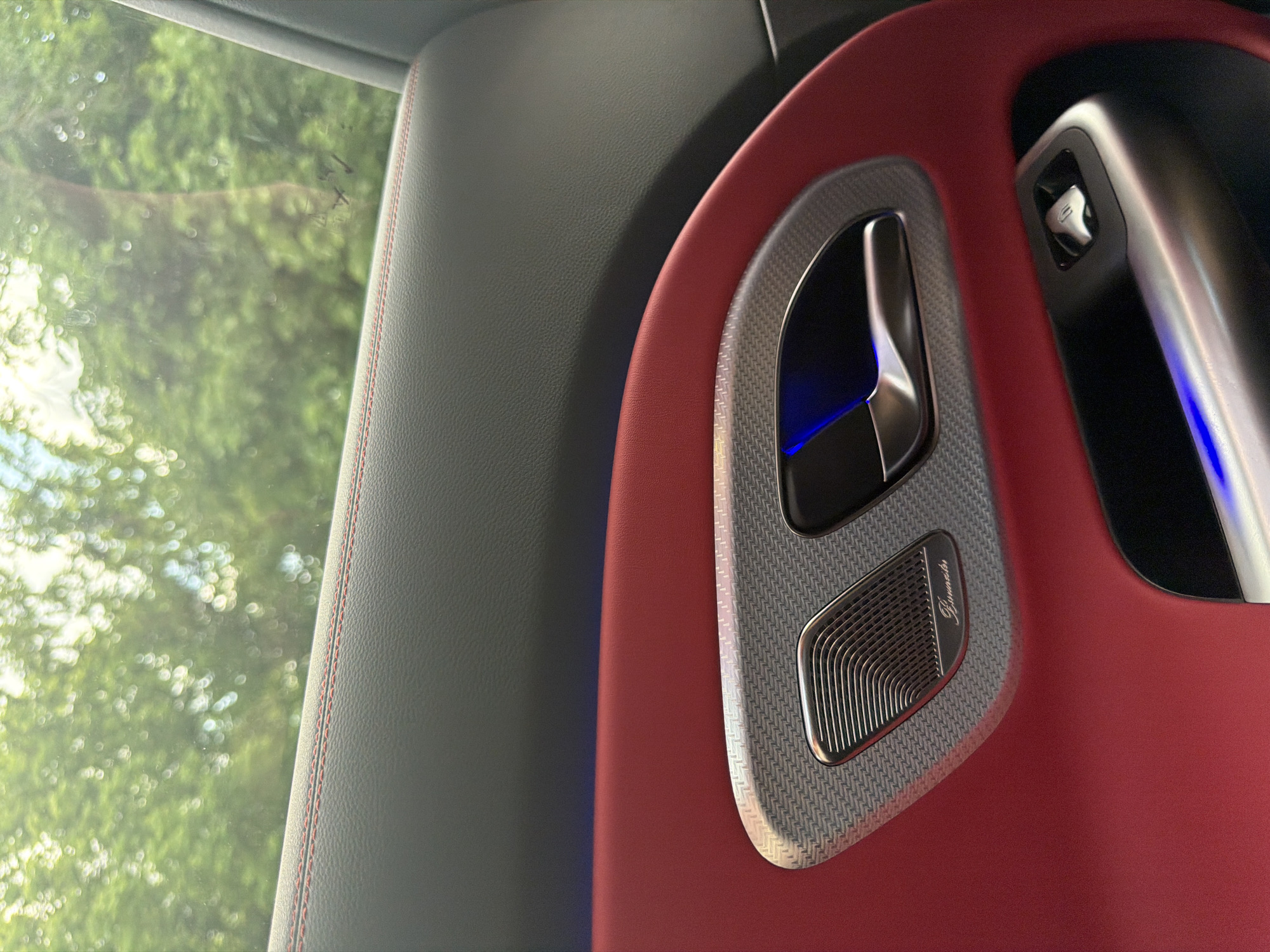 The image captures a close-up view of a luxury car's interior, specifically focusing on the front passenger side door panel. The dominant feature is a vibrant, deep red leather upholstery that contrasts with a dark grey upper door section. Red stitching accents are visible along the seams of the grey panel.
A prominent design element on the door is a speaker grille and door handle assembly. The trim surrounding these components is a dark grey, textured material, possibly carbon fiber, with a distinctive woven pattern. The door handle itself is metallic silver, sleekly integrated, and features subtle blue ambient lighting emanating from its recess. Below the door handle, a metallic speaker grille is visible, sporting the "Burmester" logo, identifying the high-end audio system.
Further right, a smaller metallic control, likely for windows or mirrors, is embedded in the dark grey trim, also showing a hint of blue illumination. Through the car window on the left, lush green foliage is visible, appearing out of focus with bokeh light effects, suggesting bright daylight conditions outside. The scene implies a stationary or slow-moving vehicle, with the focus on the intricate details and luxurious materials of the car's interior. No explicit activity or people are present.
