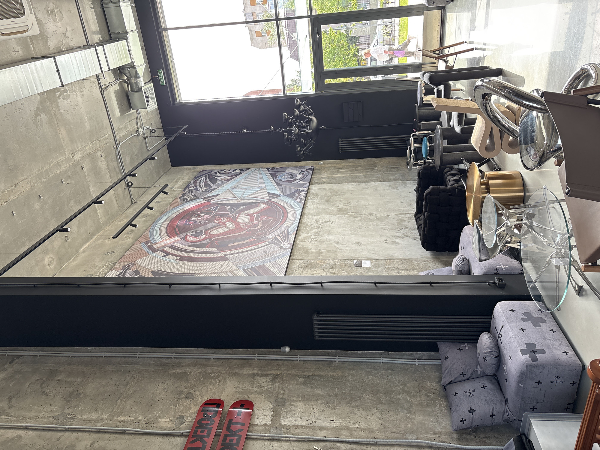 This image depicts an indoor space in what appears to be a retail or showroom setting, likely in Kazan, Russia, based on the text visible on a skateboard. The space features exposed concrete walls and ceilings, with industrial-style ventilation ducts and track lighting fixtures. A large, circular art piece dominates one wall. Below the artwork and a black structural beam, are two red skateboards leaning against the wall. The floor is also concrete.
On the right side of the image, there are various pieces of furniture and decorative items displayed, including chairs, sofas, and glass-topped tables. One sofa is gray with a cross-shaped pattern. A black, woven sofa is also visible. Natural light streams through large windows. The perspective of the image is looking upwards.