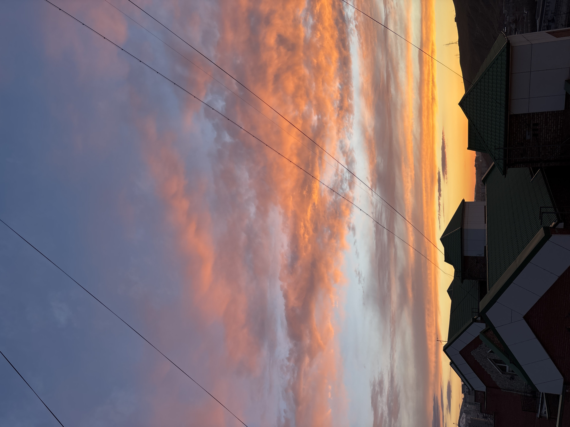 The image captures a vibrant sunset or sunrise over a city, likely Krasnoyarsk, Russia. The sky is the most prominent feature, displaying a spectacular array of colors. Streaks of orange and yellow clouds dominate the horizon, transitioning upwards into softer pinks and purples, and then to a muted blue-grey in the upper left corner. The clouds are elongated and appear to be moving, creating dynamic patterns across the sky.
Several dark utility wires run diagonally across the sky from the upper left to the lower right, adding a man-made element to the natural panorama. In the foreground, on the right side of the image, the silhouettes of multiple buildings are visible. These structures feature distinctive dark green, possibly tiled, roofs, white trim, and dark brick facades. The low light of dawn or dusk casts them in shadow, emphasizing their architectural shapes.
Beyond the buildings and directly beneath the colorful horizon, a dark, mountainous or hilly landscape is silhouetted. Faint lights or structures of a city can be discerned at the base of these distant hills. The overall scene conveys the serene beauty of twilight in an urban-natural setting. There is no visible text in the image.