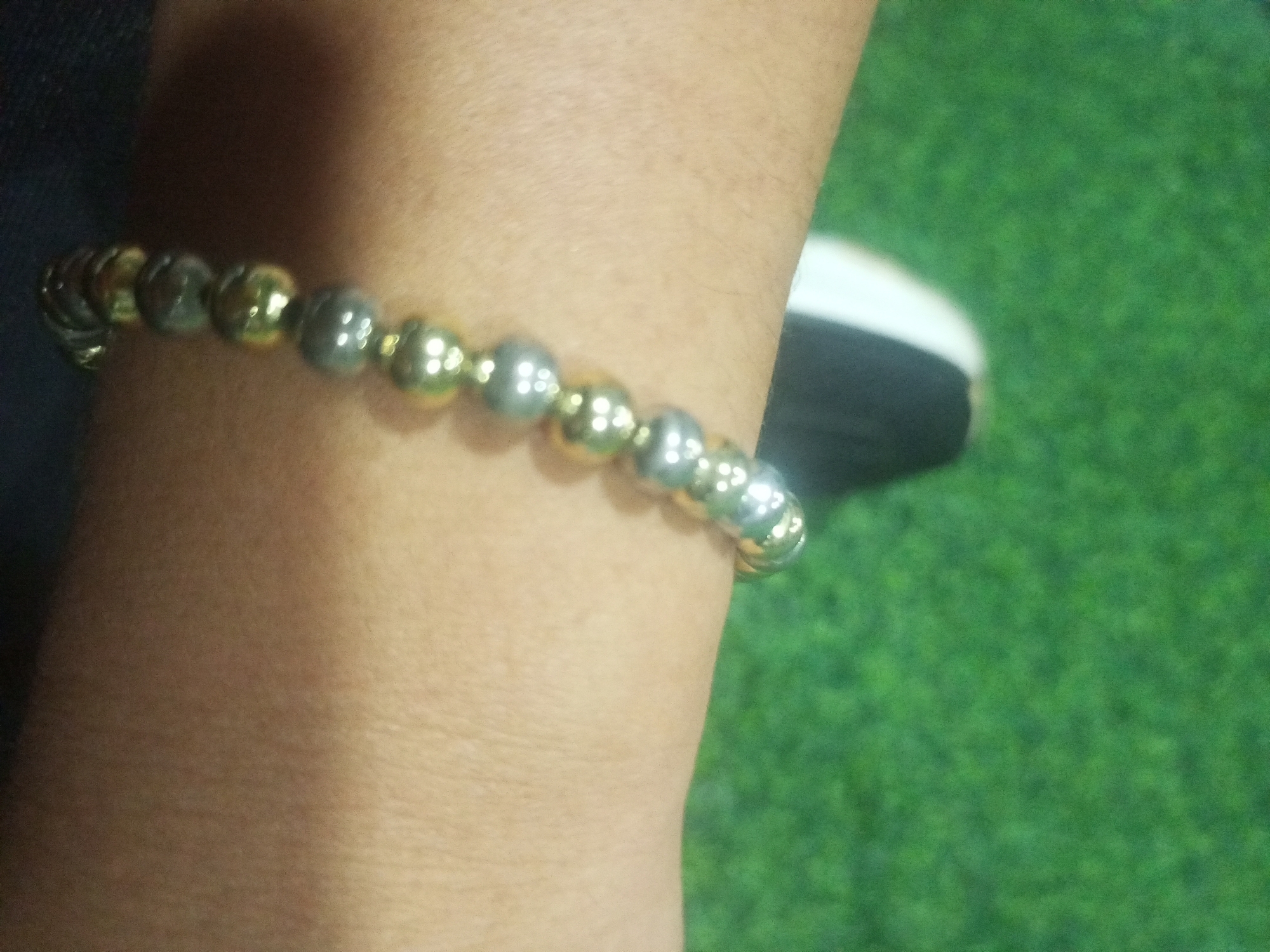 A close-up shot shows a bracelet made of alternating gold and silver beads worn around a person's wrist. The bracelet is the primary subject, with the beads appearing spherical and smooth. The wrist is light-skinned. In the blurred background, a patch of green, resembling artificial turf or grass, is visible, along with a hint of dark fabric and a white stripe, possibly from a shoe. There is no identifiable location within the city of Juan Griego, Venezuela, visible in this image.