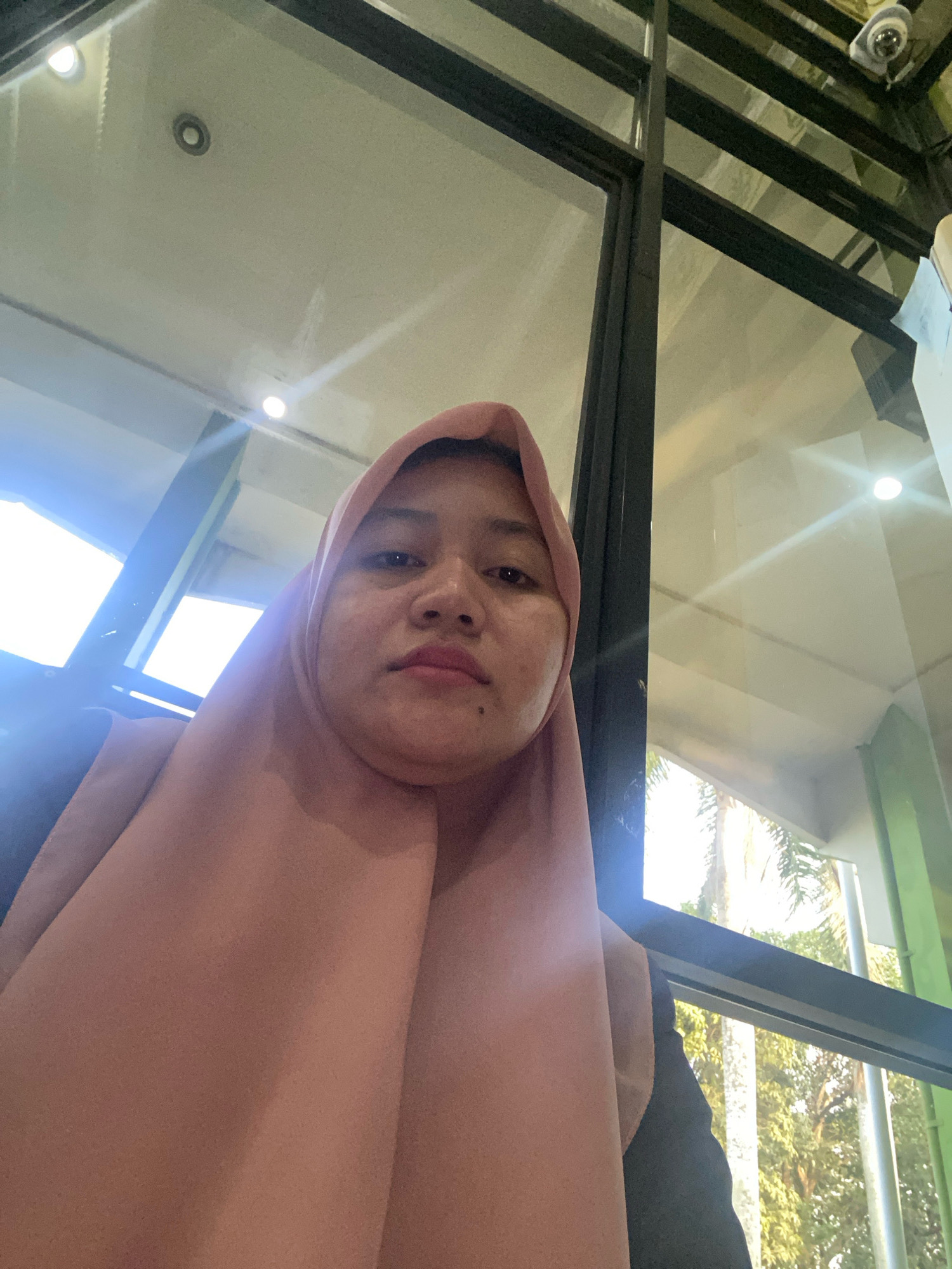 The image is a close-up, waist-up selfie of a young woman wearing a light pink hijab. She is looking directly at the camera with a neutral expression. She is indoors, seated in front of a large window with a black frame. Through the window, trees and foliage are visible, suggesting an outdoor environment. The ceiling above has lights and a security camera. The reflection on the window is not clear enough to discern specific details of the surrounding environment. The overall lighting is bright, with some lens flare present. Based on the presence of palm trees and the general architectural style, this location could be a modern building in Banjarbaru, Indonesia.