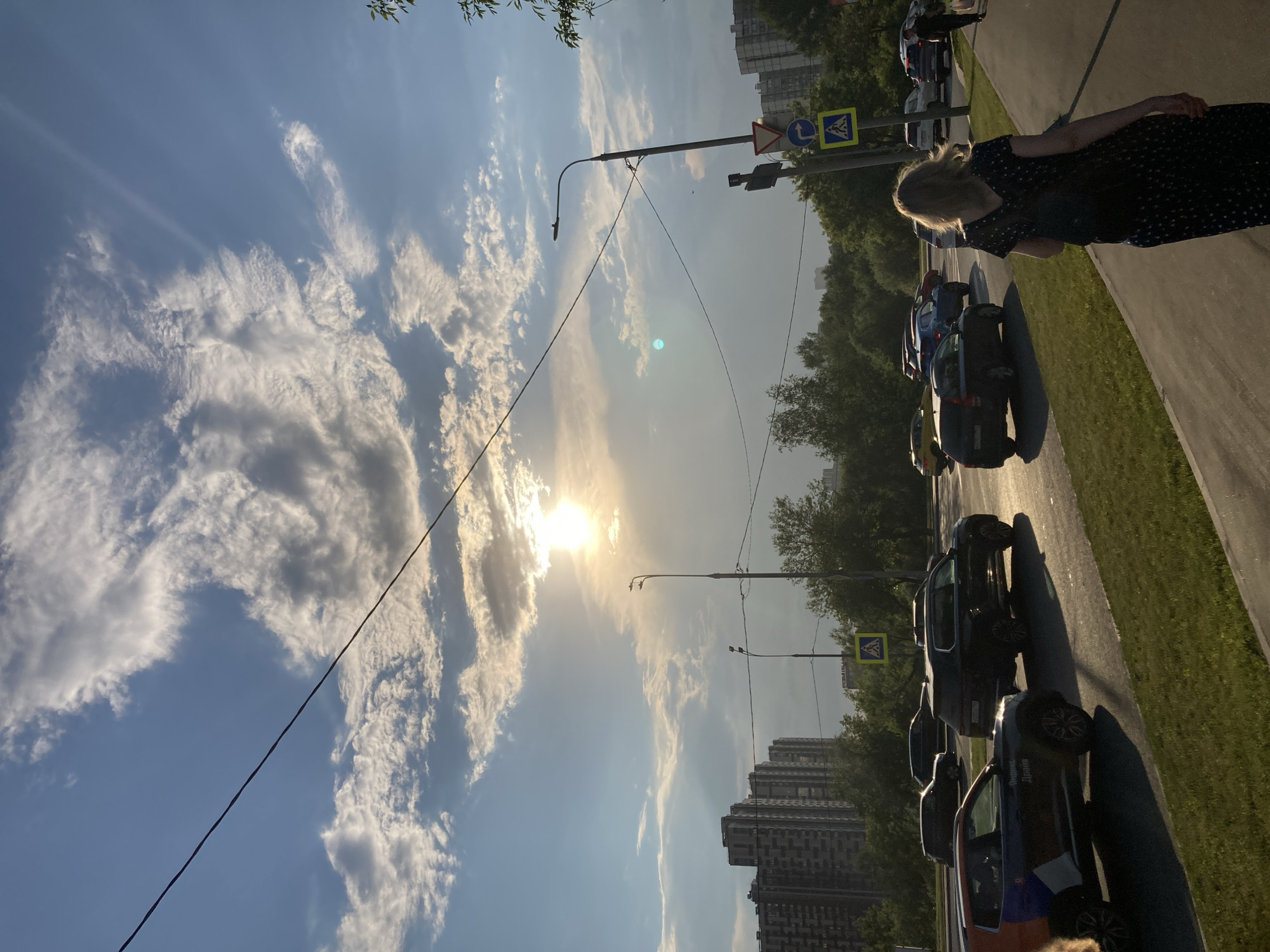 This is a street-level, eye-level shot taken from a sidewalk looking towards a multi-lane road in Moscow, Russia, during what appears to be late afternoon or early evening due to the low sun angle and long shadows. The sky is partially cloudy with bright sunlight peeking through, creating a lens flare and illuminating the clouds.
In the foreground, a woman with blonde hair, wearing a dark, patterned dress, walks away from the camera along a paved sidewalk. A dark backpack is visible on her back. The sidewalk is bordered by a grassy median.
The road features several cars. On the left side of the road, a dark-colored SUV is stopped, and to its left, another dark vehicle is in motion. Further down the road, more cars are visible, including a dark SUV and a lighter-colored sedan. A car with a distinctive orange, blue, and white livery, possibly a taxi or ride-share vehicle with "Яндекс Драйв" (Yandex Drive) written on it, is in the left lane.
Across the road, a dense line of trees separates the roadway from a backdrop of tall, multi-story residential buildings. The buildings are modern, block-style apartments. Streetlights and traffic signals are present along the roadside. A pedestrian crossing sign is visible on a pole with traffic signs, including a right-turn arrow and a yield triangle. Overhead power lines stretch across the scene. The overall impression is of a typical urban environment with active traffic and residential areas bathed in the warm light of the setting sun.