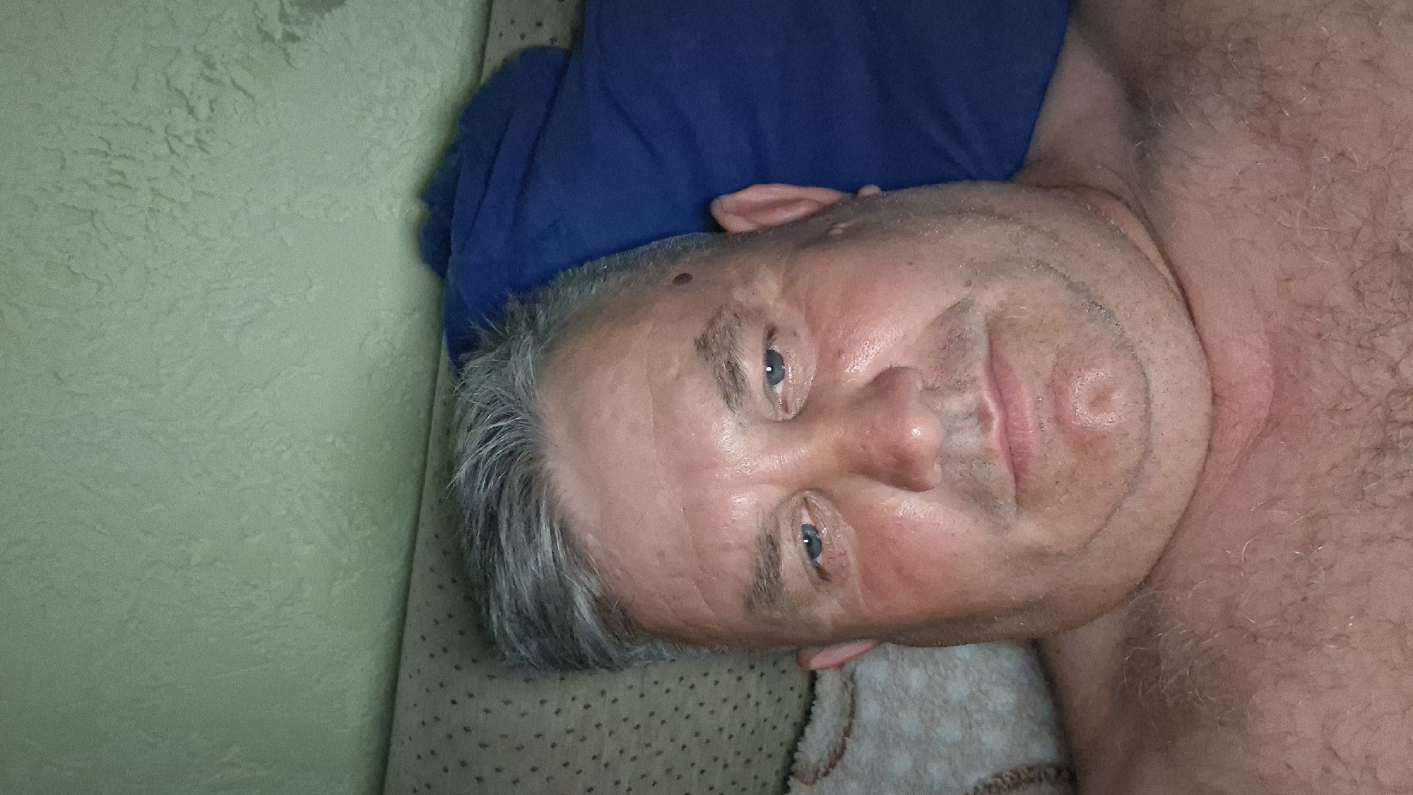 A selfie is taken from a low angle by a fair-skinned adult male with graying hair, who is lying down. He is shirtless, showing a hairy chest, and resting his head on a blue pillow. The background consists of a textured, light green wall and a portion of a patterned cushion. The man's face is the primary focus, with his blue eyes looking directly at the camera. He has some facial stubble and a mole on his left temple. The overall lighting is dim, suggesting an indoor setting, possibly a bedroom. There is no visible indication of the location being in the city of Voinova, Moldova.