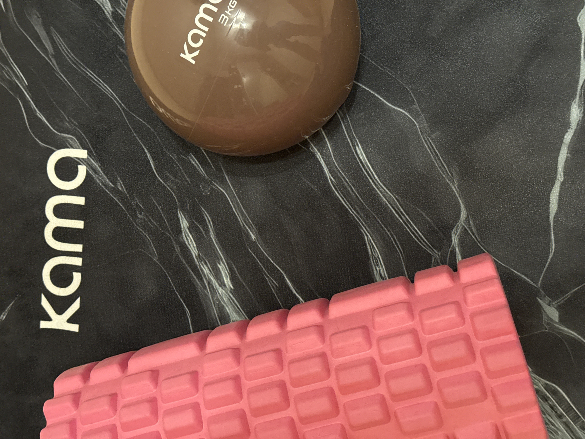 The image shows a top-down, close-up view of fitness equipment on a dark, marble-patterned mat. On the left side of the frame is a portion of a bright pink foam roller with a textured surface composed of rectangular protrusions. To the right of the foam roller is a brown, spherical weight, possibly a medicine ball. The word "kama" is visible in white lettering on both the mat and the weight. Below "kama" on the weight, "3KG" is also visible. The background is a black mat with swirling white lines resembling marble veining. There are no people or any indication of a specific location within a city.