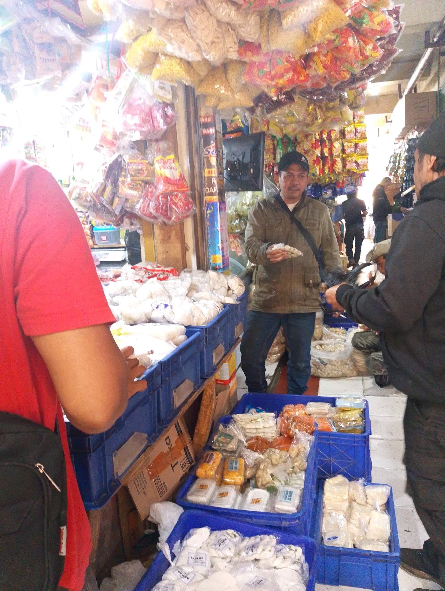 The image depicts a street market scene in Lembang, Indonesia. In the foreground, a person wearing a red shirt with a black bag slung over their shoulder is partially visible on the left. In the center, a man in a green jacket and black baseball cap is holding a small bag of snacks. He is standing in front of a stall laden with various packaged food items, including chips and snacks hanging from the ceiling. To the right, another person in a black jacket appears to be interacting with the man. The stall is filled with blue crates containing more packaged food items, as well as some loose items in plastic bags. The market appears to be bustling with activity, with other shoppers and vendors visible in the background. The overall atmosphere suggests a lively commercial environment.