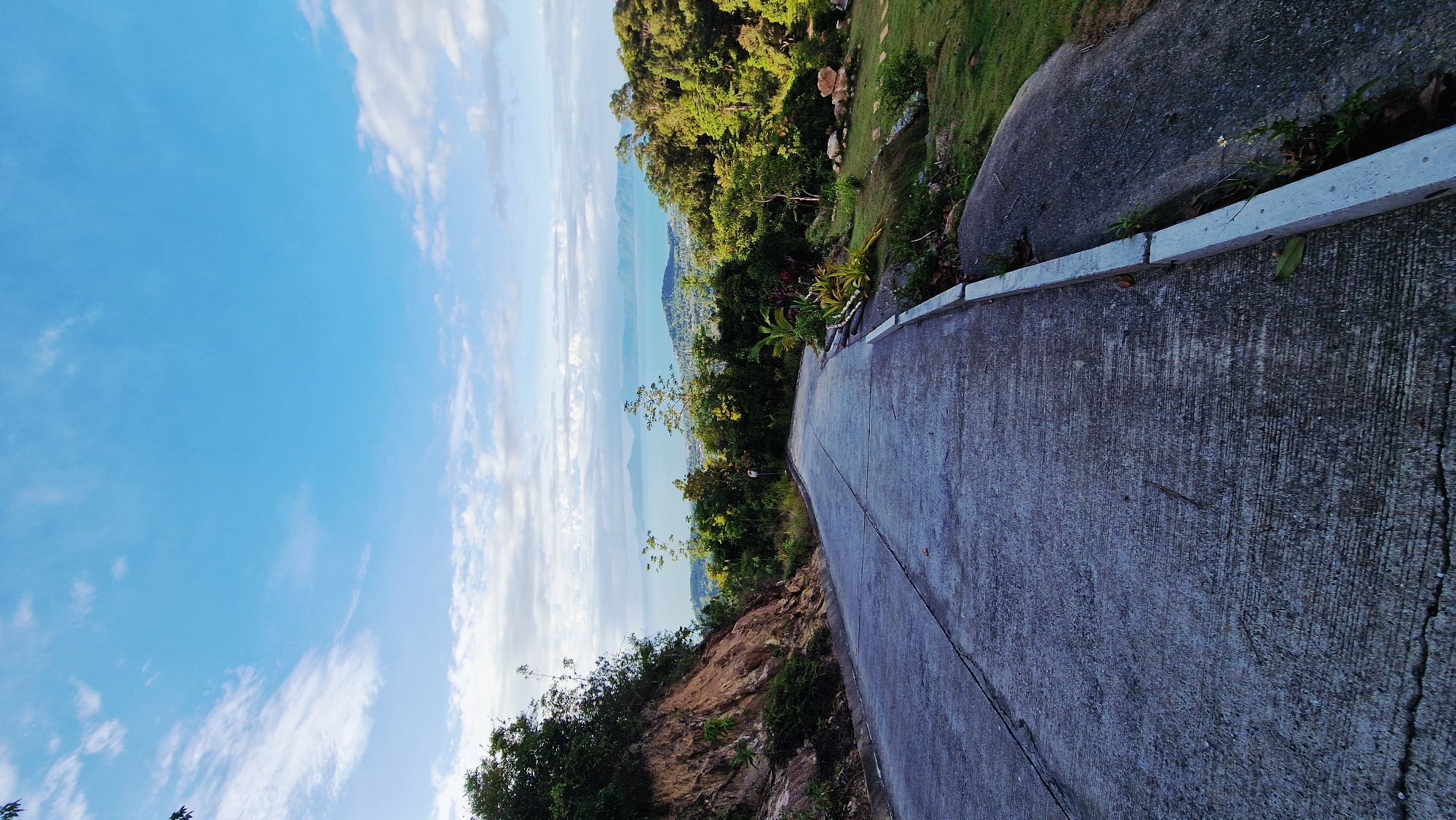 The image depicts a concrete road curving to the right, leading towards a scenic overlook. On the left, a steep, rocky embankment covered with vegetation rises alongside the road. To the right of the road, a grassy slope is dotted with rocks and lush green trees, with some tropical plants visible near the curb. In the distance, a body of water, likely the sea, stretches across the horizon, with islands and a hazy coastline visible under a bright blue sky with scattered white clouds. The overall scene suggests a rural or semi-rural location, possibly a viewpoint or a road leading to one in a tropical region, consistent with the city of Baan Tai, Thailand. There are no people or animals visible in the image.