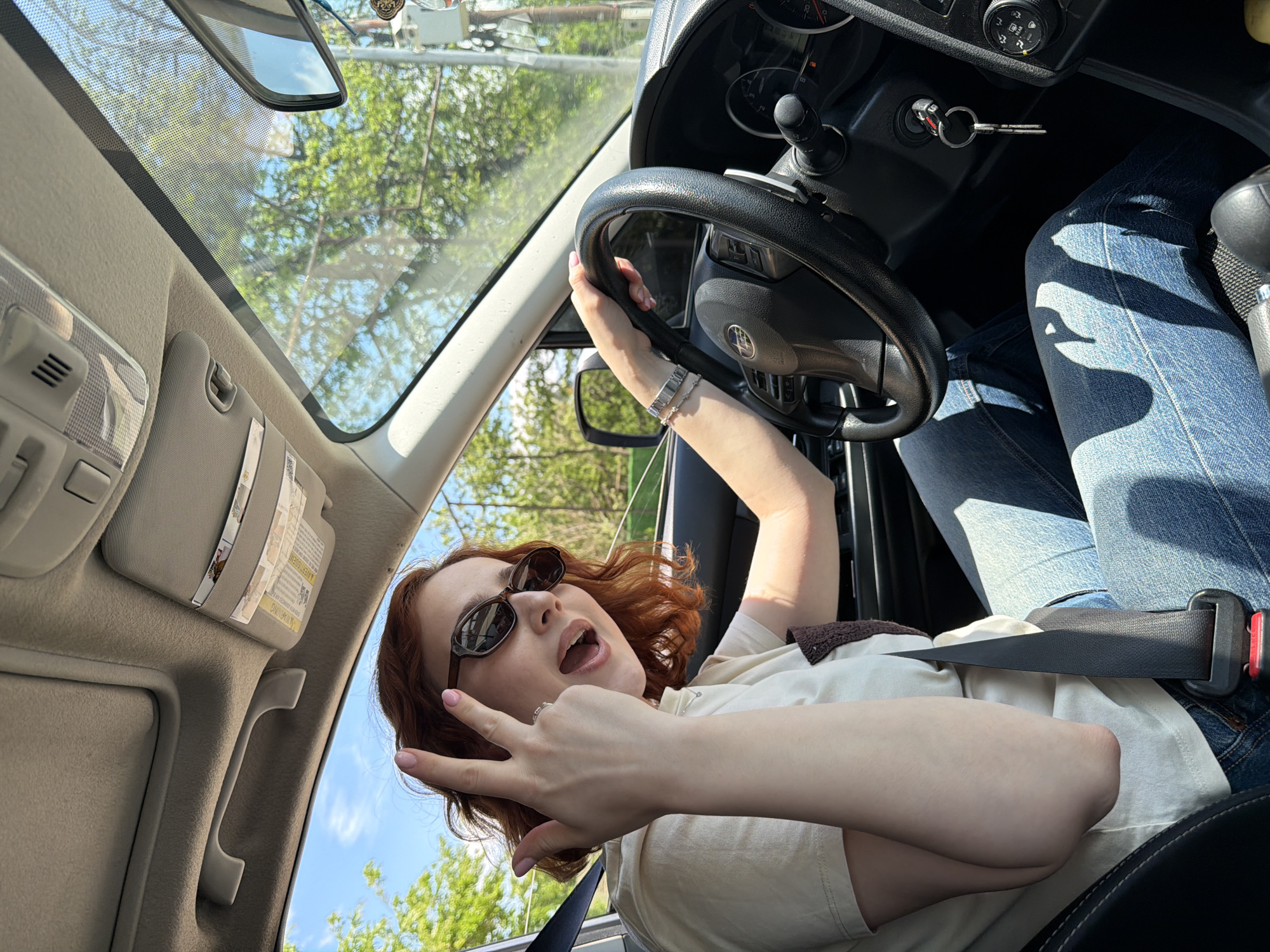 The image is taken from inside the cabin of a car. A young woman with auburn, wavy hair, wearing sunglasses, a light-colored shirt, and blue jeans, is sitting behind the steering wheel. She is making a hand gesture, specifically a "peace" or "victory" sign, towards the camera. Her seatbelt is fastened. The car keys are in the ignition. Outside the car, trees and the sky are visible through the windshield. Based on architectural hints within the image, it is potentially located in Tbilisi, Georgia.