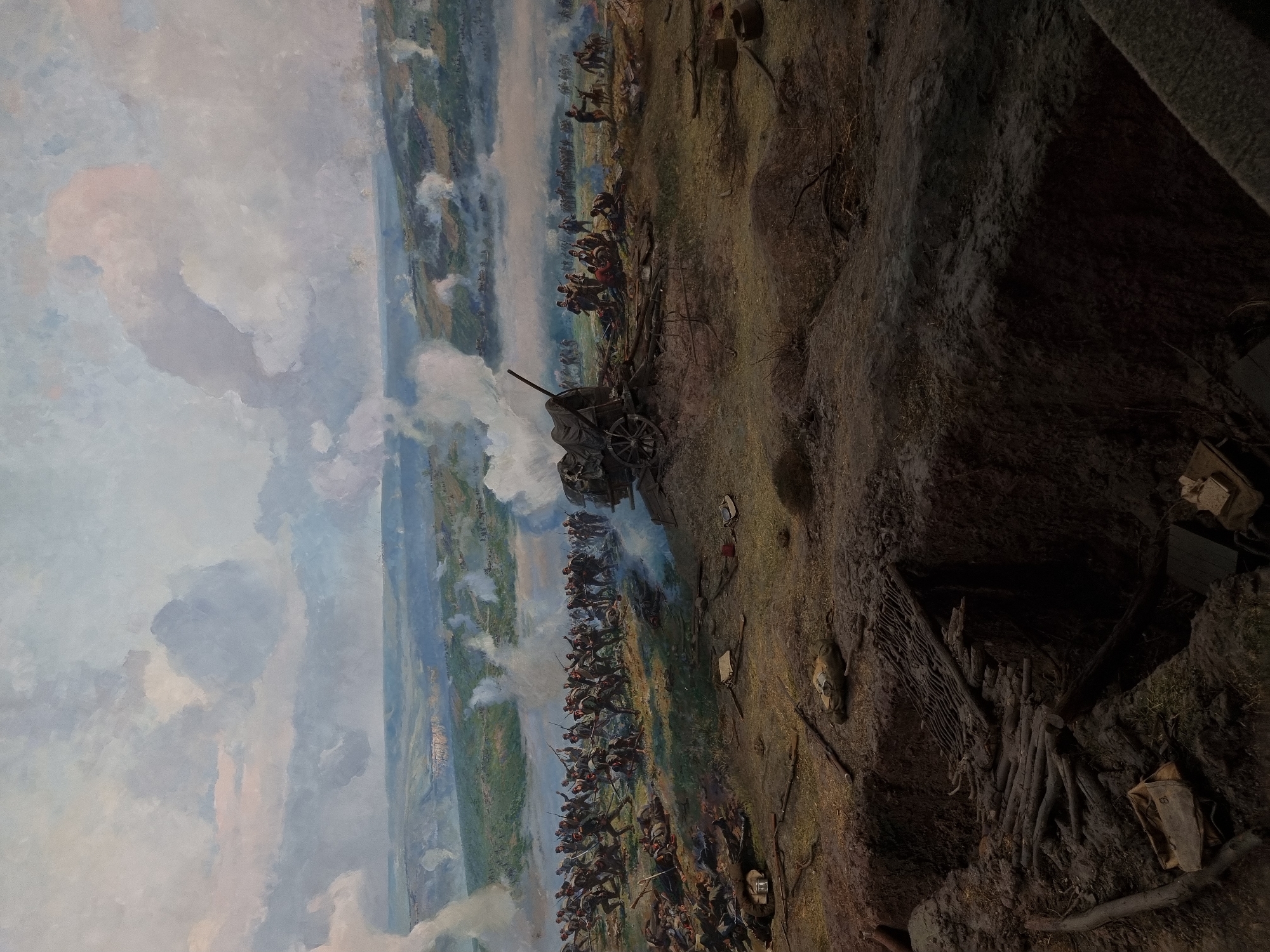 The image depicts a panoramic painting of the Siege of Pleven, a major battle in the Russo-Turkish War of 1877-1878, likely located in the city of Pleven, Bulgaria, where a museum dedicated to the event exists. The painting showcases a vast battlefield with various elements:
* **Environment:** A war-torn landscape, with rolling hills, vegetation, and a sky filled with smoke from artillery fire.
* **People:** Numerous soldiers engaged in combat, with visible ranks and formations.
* **Objects:** Artillery pieces (cannons), fortifications, wagons, and other military equipment are present.
* **Actions:** Soldiers are depicted firing rifles, advancing, and engaging in close-quarters combat.
* **Interactions:** The painting portrays a large-scale military confrontation between opposing forces.
* **Foreground:** A trench system and other defensive structures.