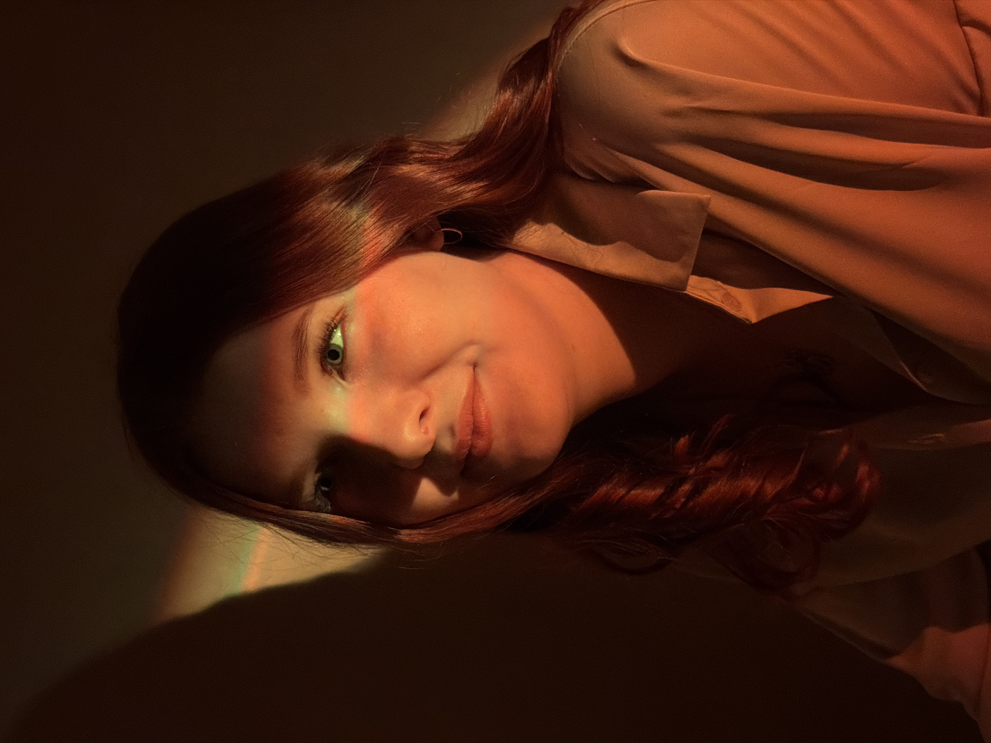 A close-up, medium shot depicts a young woman with fair skin and red hair, tilted at an angle. She is wearing a tan-colored blouse. Her eyes are open, and she has a slight smile. A warm, orange-toned light illuminates her face, with a rainbow-colored line accentuating the left side of her face. The background is a gradient of dark brown to light tan. The location is unknown and there is no indication of the city of Chaikovsky, Russia in the image.