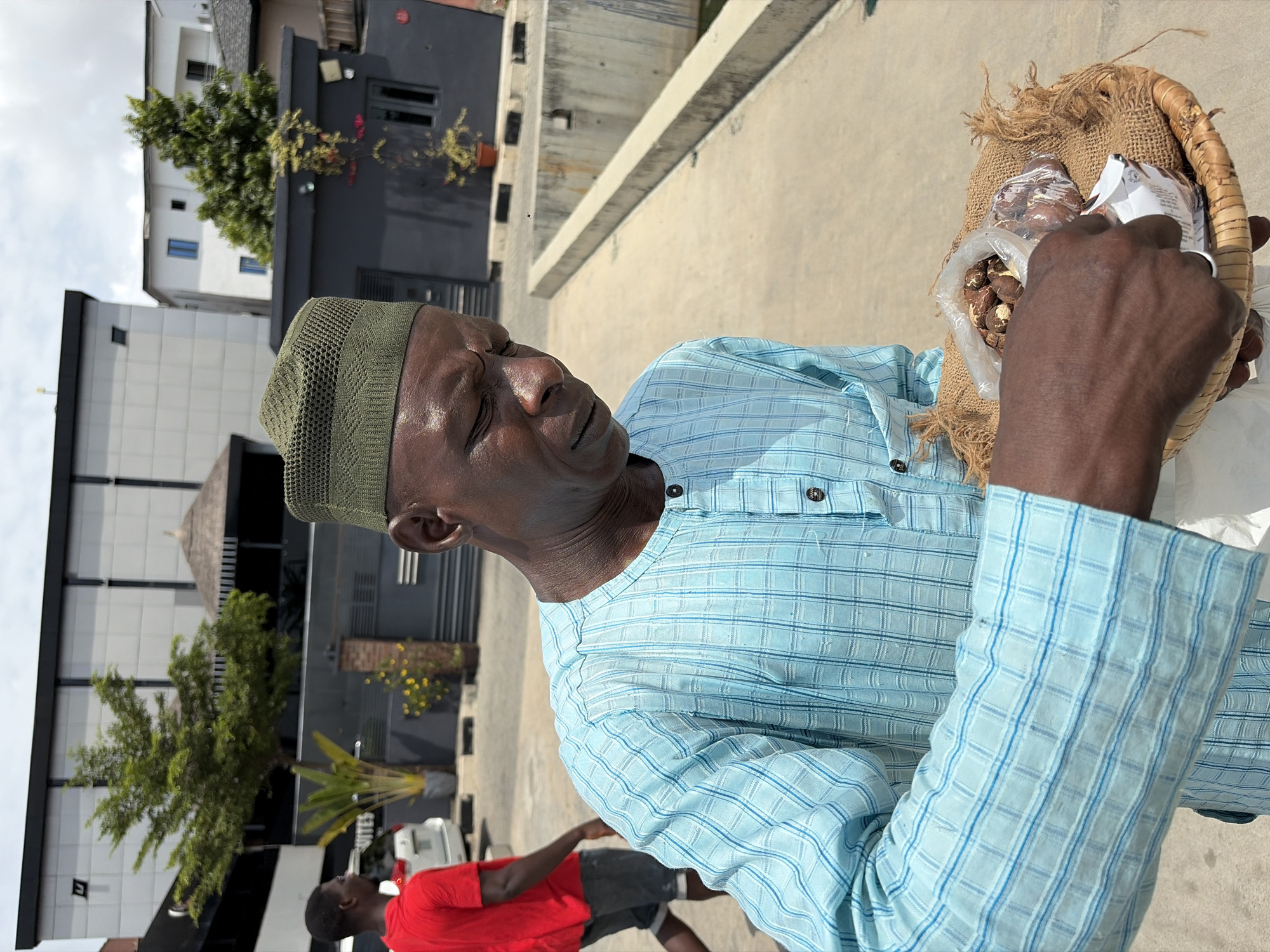 An adult Black male wearing a green knitted cap and a long-sleeved light blue checkered shirt holds a small woven basket in his right hand. The basket has a burlap liner. The basket contains a clear plastic bag filled with brown nuts and white paper packaging. The man looks down toward the basket.
In the background, a person wearing a red shirt and dark shorts walks away in the lower left corner. Behind the man, there are multi-story buildings and trees. A concrete barrier separates the man from the building and a parking lot behind the building has a white car.
The scene is likely in Igboefon, Nigeria, based on the request.