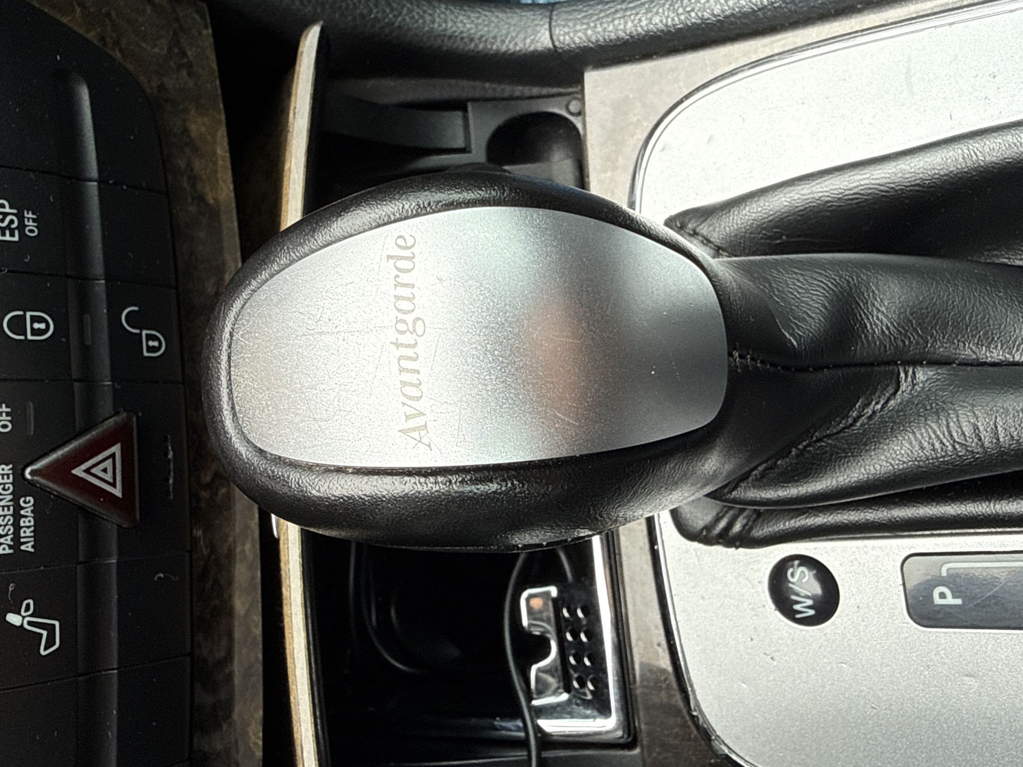 The image displays the interior of a vehicle located in Stara Zagora, Bulgaria, focusing on the center console and automatic transmission gear selector. The gear selector knob is black with a brushed silver metallic insert prominently featuring the embossed text "Avantgarde." The shaft of the selector is covered in textured black material, likely leather, with visible stitching. Below the knob, a circular black button labeled "W/S" (Winter/Sport mode) is visible, alongside a "P" (Park) indicator, confirming the vehicle is in Park.
Above the gear selector, a row of black control buttons is visible against a dark wood-grain trim. These buttons include, from left to right: a partially visible heated seat icon, a "PASSENGER AIRBAG OFF" button, a red triangular hazard warning light button, a door lock icon (closed padlock) button, an "ESP OFF" button, and a door unlock icon (open padlock) button.
To the left and slightly forward of the gear selector, a recessed compartment is visible, containing a silver-colored metallic component with multiple small rectangular openings and circular indentations. A black cable extends into this compartment. The surrounding trim is silver-colored. No individuals, actions, or specific interactions are depicted. The scene is static, illuminated by ambient light.