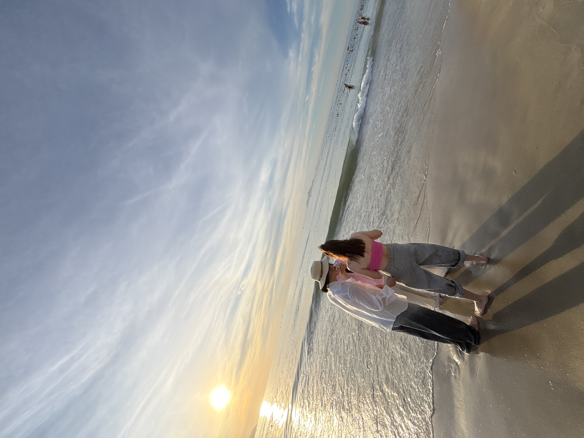 Two individuals are standing on the shore of a beach in Da Nang, Vietnam, facing the ocean. The person on the left is wearing a white long-sleeved shirt, dark pants, and a straw hat. The person on the right is wearing a pink halter top and striped pants. They appear to be interacting, with the person on the left leaning towards the person on the right. The sun is setting or rising, casting a warm glow on the water and sky, with wispy clouds stretching across the horizon. In the distance, other beachgoers are visible in the water. The tide is coming in, with small waves lapping at the individuals' feet.