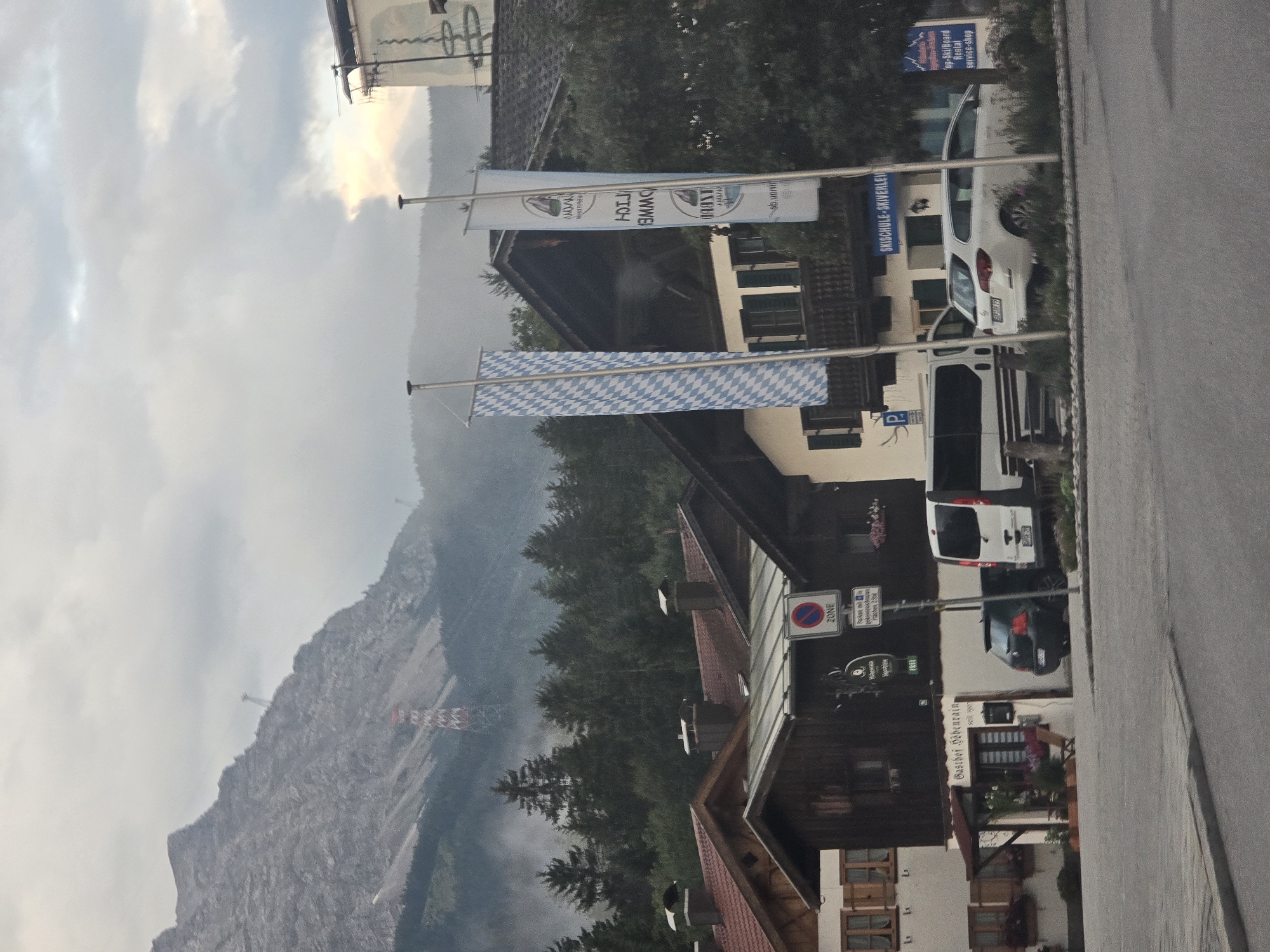 The image depicts a street scene in Grainau, Germany, with a prominent mountain in the background. The mountain is rugged and rocky, with a cable car visible ascending its slope, and a tall, red and white pylon structure situated partway up. The sky is overcast with scattered clouds, and a hazy mist hangs over the treeline that separates the mountain from the town.
In the foreground, several buildings line the street. The architecture suggests a Bavarian style, with dark wood accents, red tile roofs, and dormer windows. One building has a sign above the entrance that reads "Gasthof Höhenrain seit 1907," indicating it is an inn or guesthouse. Another building has a sign that says "SKISCHULE - SKIVERLEIH," meaning ski school and ski rental.
Several vehicles are parked along the street, including a dark-colored hatchback and a white van. Two flags are prominently displayed on poles in front of the buildings; one is a Bavarian flag with a blue and white diamond pattern, and the other is a white banner with blue lettering that appears to be an advertisement. Traffic signs are also visible, including a "ZONE" sign and a parking sign.
The overall impression is a quaint, alpine village setting, likely a tourist destination given the presence of the ski school and the mountainous backdrop. The lighting suggests it is either early morning or late afternoon due to the soft, diffused light and the presence of mist. No people are clearly visible in the image.