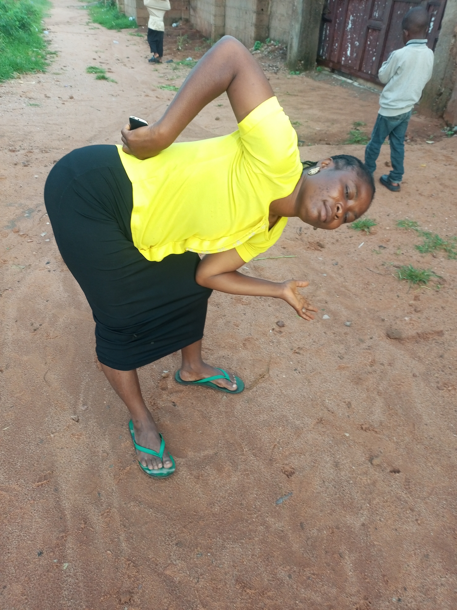 The image depicts an outdoor scene on a dirt path. In the foreground, a woman wearing a bright yellow t-shirt and a long black skirt is bent over, holding a black mobile phone. She is wearing green flip-flops. Her expression is directed upwards and to the right. In the middle ground, a young boy in a light-colored long-sleeved shirt and blue jeans stands facing away from the camera. In the background, another person, possibly a child, is partially visible in a light-colored top and dark pants, walking away. The environment consists of a dirt path with sparse green vegetation along the edges and a fence with dark wooden panels to the right. The overall impression is of a casual, everyday moment in a semi-urban or rural setting in Tempure, Nigeria.