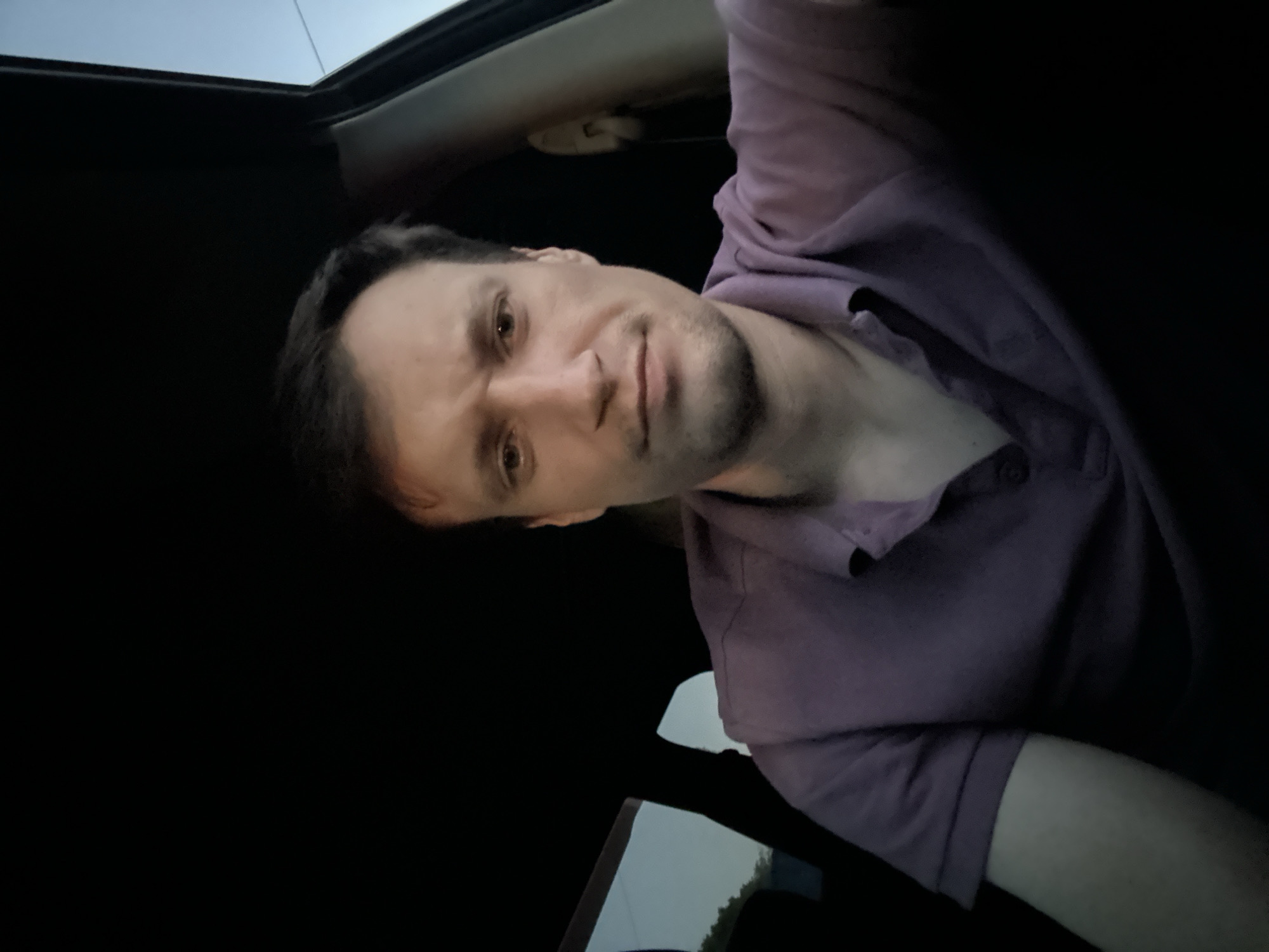 A man is sitting in the driver's seat of a car. He is looking directly at the camera and has a neutral expression. The interior of the car is dark, with the only light source coming from the outside, suggesting it is either dawn or dusk. The man is wearing a purple polo shirt. The background outside the car window shows a pale sky and some greenery. The scene appears to be outdoors in an unidentifiable location.