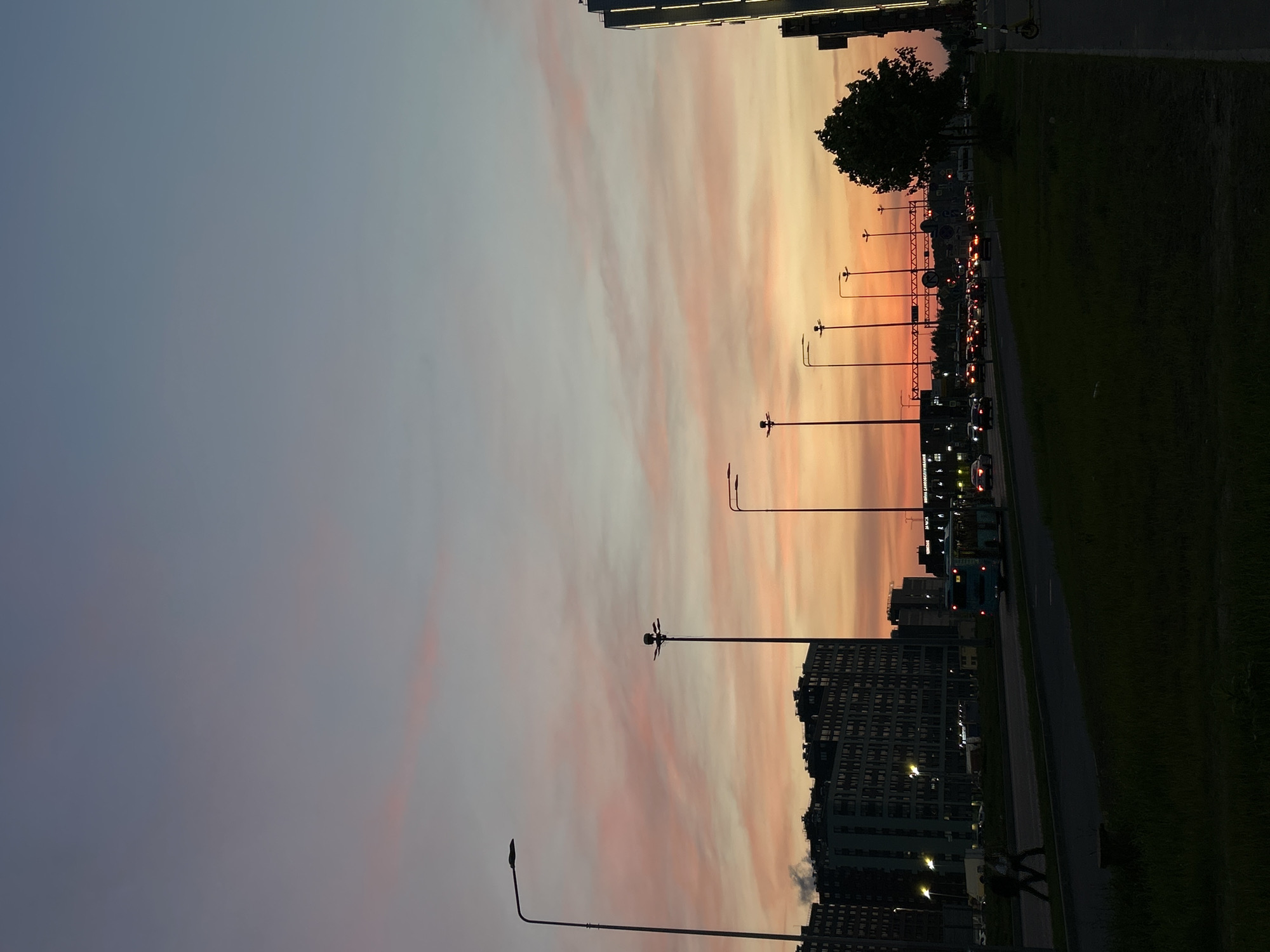 The image captures a street scene in Saint Petersburg, Russia, at dusk. The sky displays a gradient of colors, transitioning from gray on the left to orange and pink towards the horizon. The scene includes buildings, streetlights, a road, a grassy area, and cars. On the right side, a road runs parallel to a green grassy area. People can be seen walking on the sidewalk. Cars are driving along the road, some with their headlights on. In the background, a row of streetlights lines the road. A large tree is visible, silhouetted against the sky. Several buildings, including apartment blocks with multiple windows, are situated along the street.