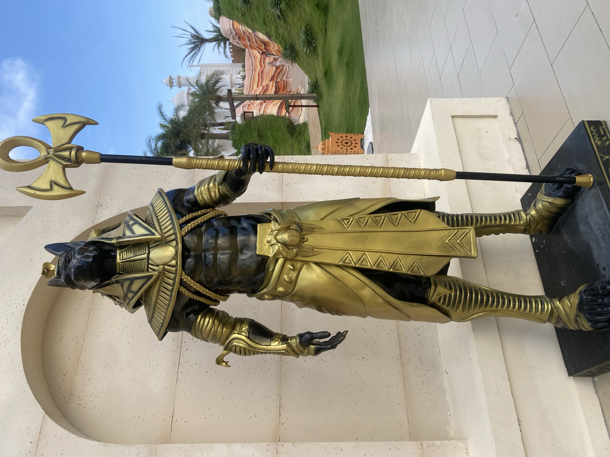 A statue of Anubis, the Egyptian god of the dead, stands in Mũi Né, Vietnam. The statue is predominantly gold and black, depicting Anubis in traditional garb, holding a staff with an Ankh at the top. In the background are white buildings, palm trees, and landscaped greenery. The environment includes a bright blue sky and a tiled walkway where the statue is placed.