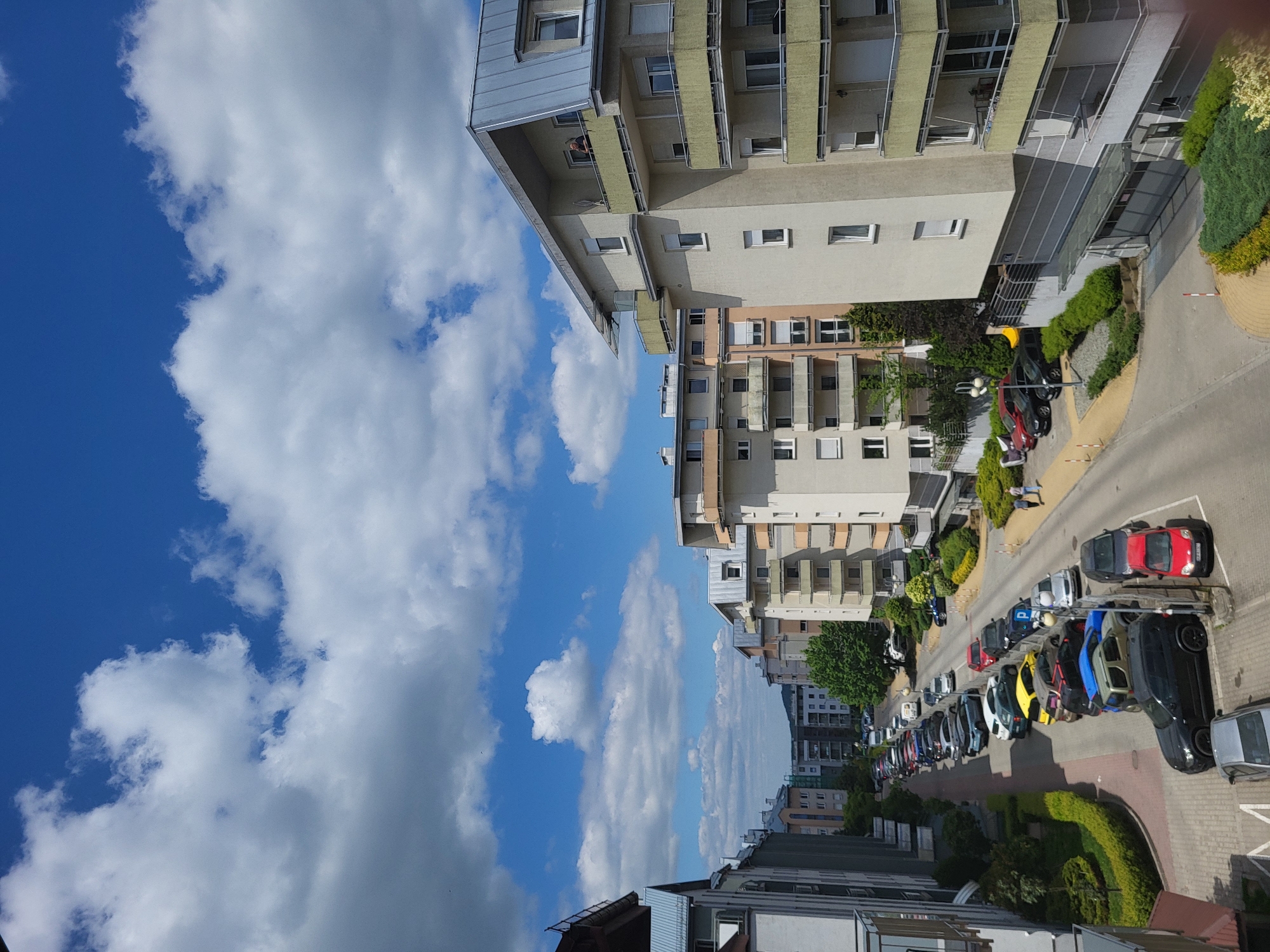 Here is a description of the image:
The image shows a street scene with tall buildings on either side. The sky is blue with white clouds. A row of parked cars lines the street, with colors including red, blue, yellow, and black. Trees and greenery are visible along the sidewalks. The location appears to be a residential area in a city, though the specific location is unknown.