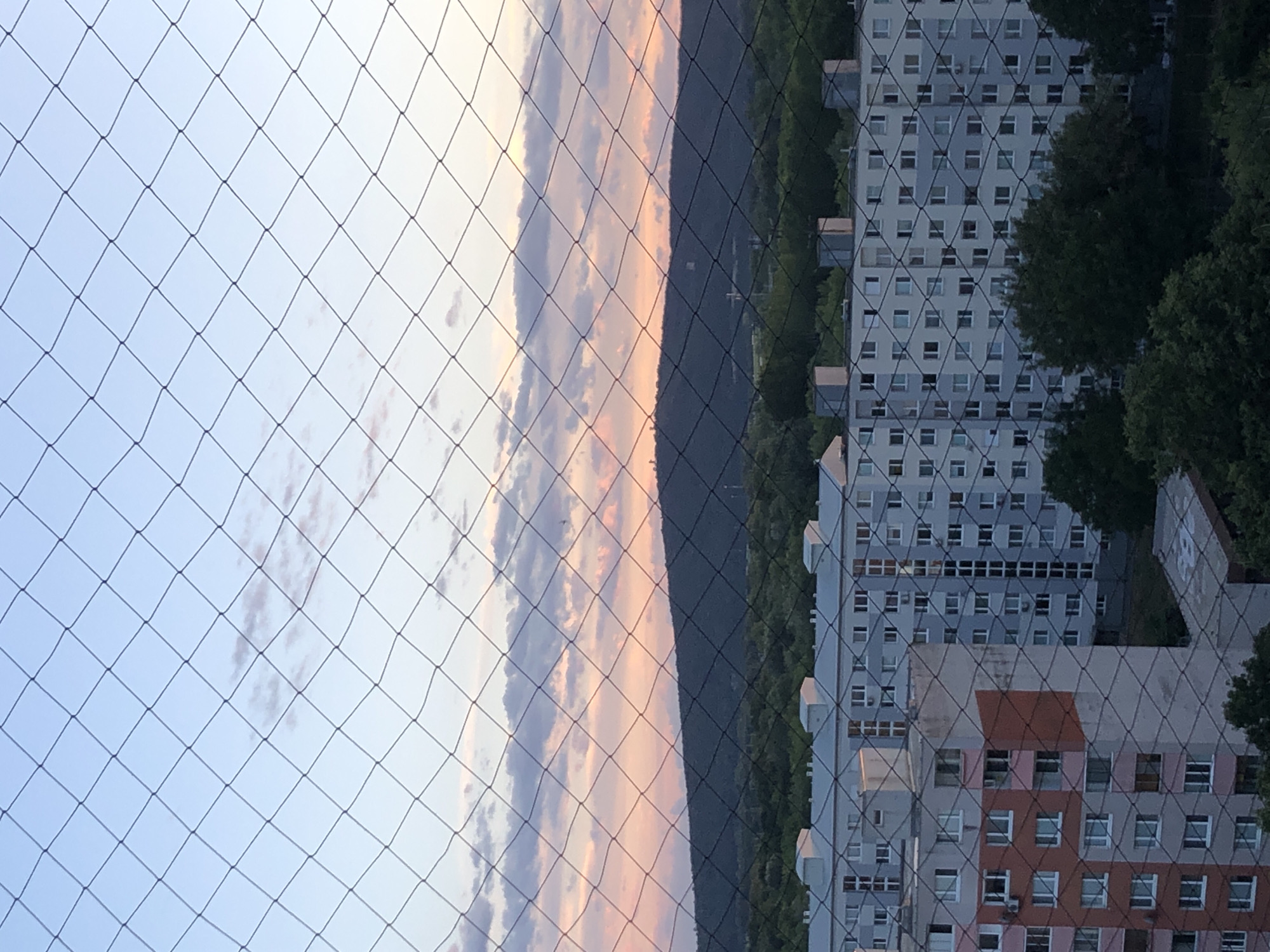 The image captures a sunset view over Bratislava, Slovakia, as seen through a protective net. In the foreground, a black net with diamond-shaped openings overlays the scene. Below the sky, a large, dark hill dominates the middle ground, with green trees covering its lower slopes. Several buildings, characteristic of Soviet-era architecture, are visible in the foreground. One building on the left is taller and features a distinctive pattern of reddish-orange and grey sections, with numerous windows. To its right, a long, white apartment block stretches across the frame, also covered in the netting, with many windows. The sky above is a soft blend of blues, pinks, and oranges, indicating the twilight hour, with scattered clouds. The overall impression is an urban landscape bathed in the gentle light of sunset.