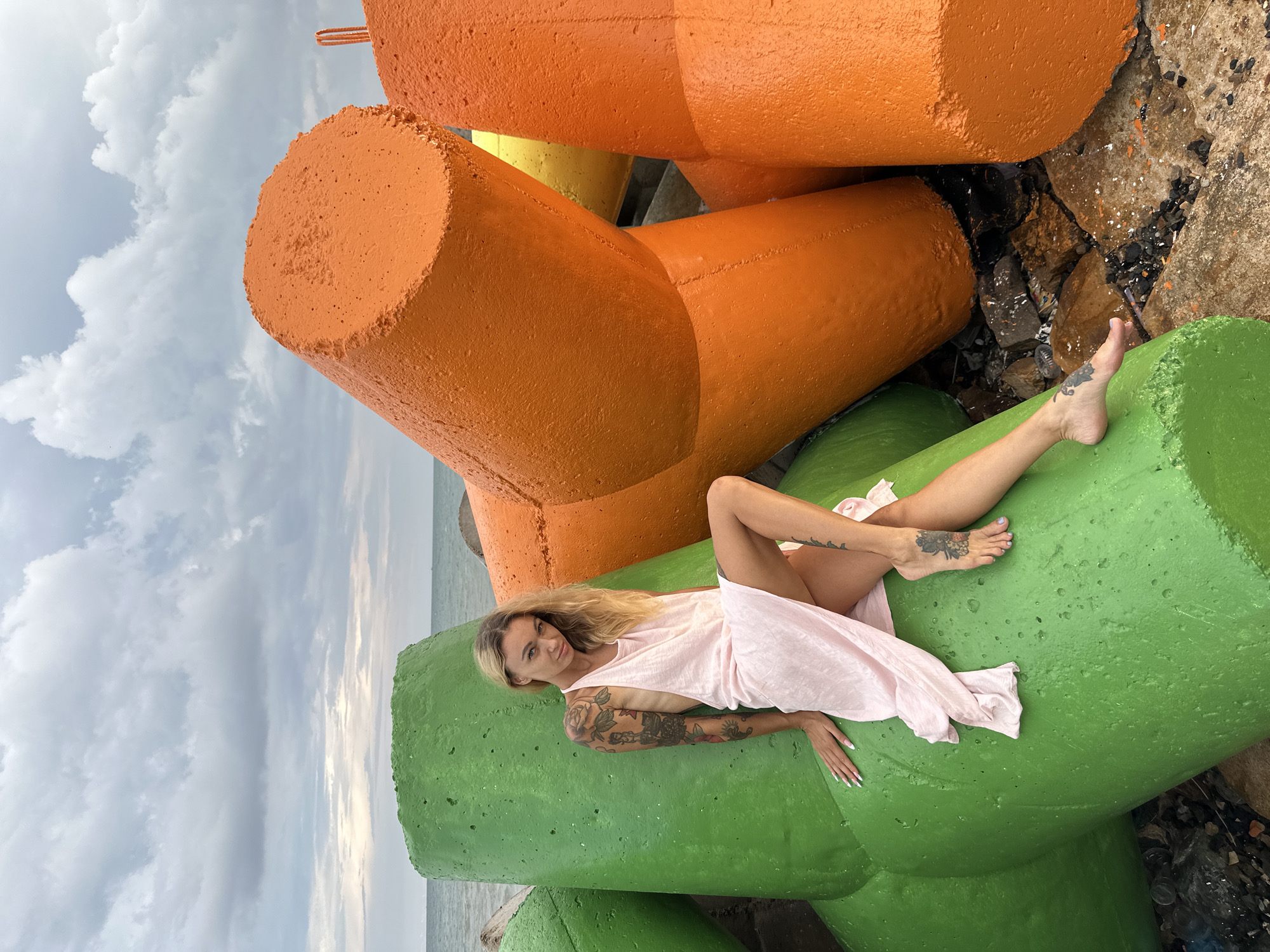 The image displays a woman posing on large, brightly colored concrete tetrapods at a coastal location. The woman, with long blonde hair, lies on her left side on a vibrant green tetrapod in the foreground, looking directly at the camera. She is dressed in a flowing, light pink sleeveless dress. Visible tattoos adorn both of her arms and her right foot. Her expression is calm and relaxed, conveying a soft smile.
Behind her, several large, angular concrete tetrapods are arranged, predominantly painted in a bright orange hue. A smaller section of a yellow tetrapod is also visible between the orange structures. The tetrapods exhibit a rough, textured surface. To the right, beyond these structures, the sea is visible under a cloudy, overcast sky, suggesting a daytime scene, likely late morning or afternoon. The ground between the tetrapods consists of natural rocks and scattered debris, marked with splashes of colorful paint. No text is visible in the image. The scene takes place in a coastal environment, consistent with Phu Thuy Ward, Vietnam.