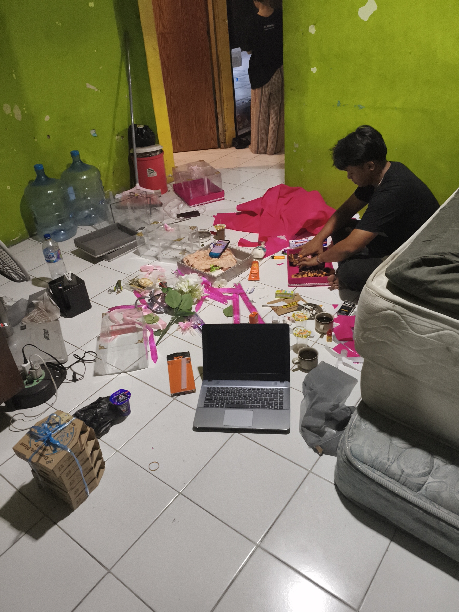 In a room with lime green walls, a young man in a black t-shirt sits on the floor, focused on a pink box filled with small items, possibly makeup or crafts. He is surrounded by a variety of objects, suggesting a cluttered or in-progress project. To his left, a laptop is open on the floor, its screen dark. Various boxes, bottles of water, and decorative items, including pink fabric and flowers, are scattered around the space. Two mugs sit near the man, possibly containing beverages. In the background, another person, a woman wearing a black top and light-colored patterned pants, stands near a doorway leading to another room. The scene appears to be indoors, possibly a home or a workshop, with tiled flooring. The lighting is dim, suggesting it might be evening or a poorly lit room. Some visible text on a pizza box reads "Domino's Pizza". The overall impression is one of a work in progress, with a sense of focus from the young man amidst the disarray. The setting appears to be in Depok, Indonesia, based on the provided location context.