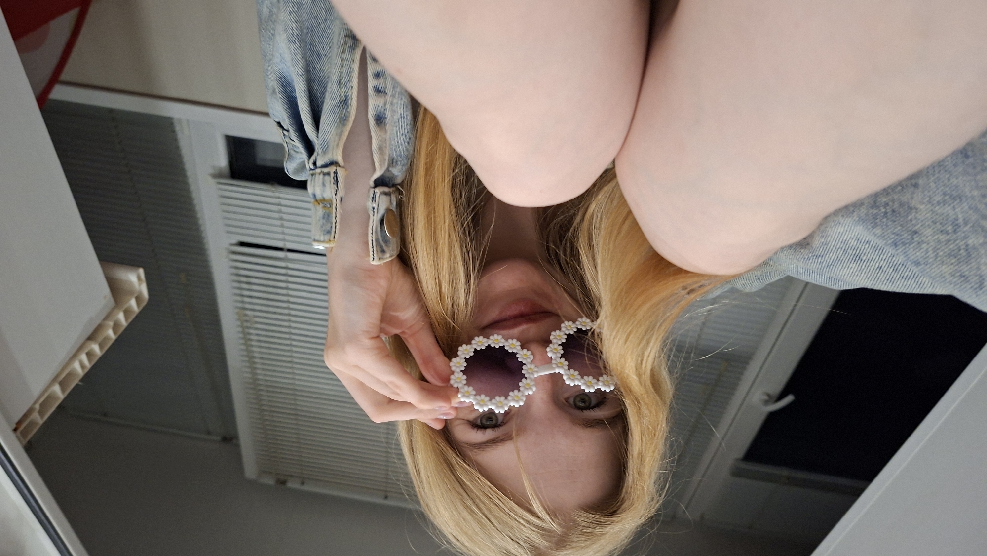 A young, fair-skinned woman with long, blonde hair is shown from the chest up, looking at the camera. She is wearing oversized, novelty sunglasses decorated with small white flowers. Her right hand is holding the sunglasses up to her face. She appears to be wearing a denim jacket, with the cuff visible on her right arm. The background consists of what appears to be blinds on a window, and a white cabinet. The lighting suggests an indoor setting.