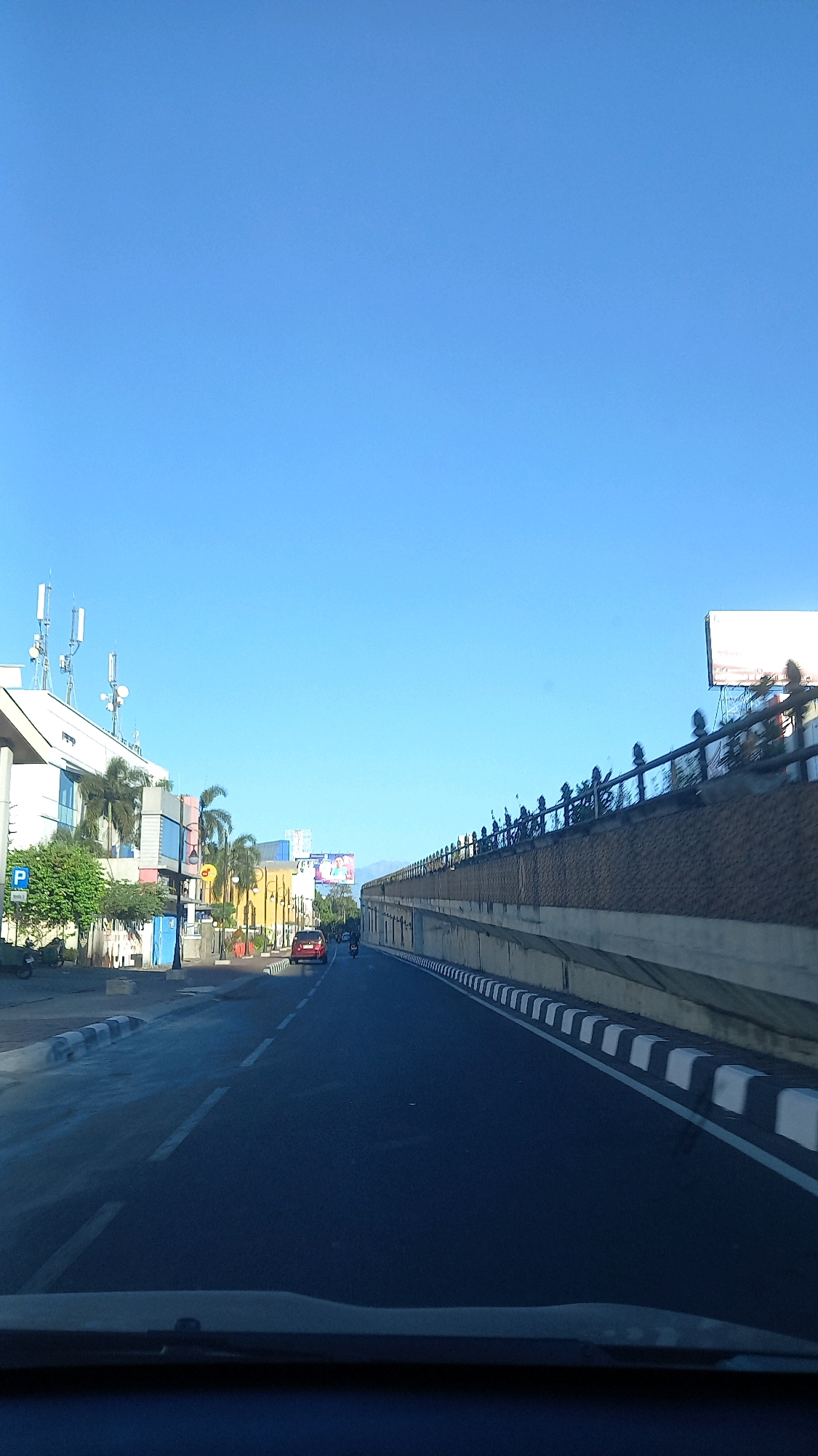 The image captures a view of a road in Surakarta, Indonesia, likely during daytime with clear skies. The road appears to be a multi-lane street with white markings. On the left side of the road, there are buildings, including one with multiple cellular antennas on its roof. There are also palm trees and a parking sign visible. Further down the road, a red car and a motorcycle are in motion. On the right side of the road, there is a raised concrete structure with a decorative fence on top, possibly an overpass or a retaining wall. A large billboard is situated atop this structure. The scene suggests an urban environment with ongoing traffic.