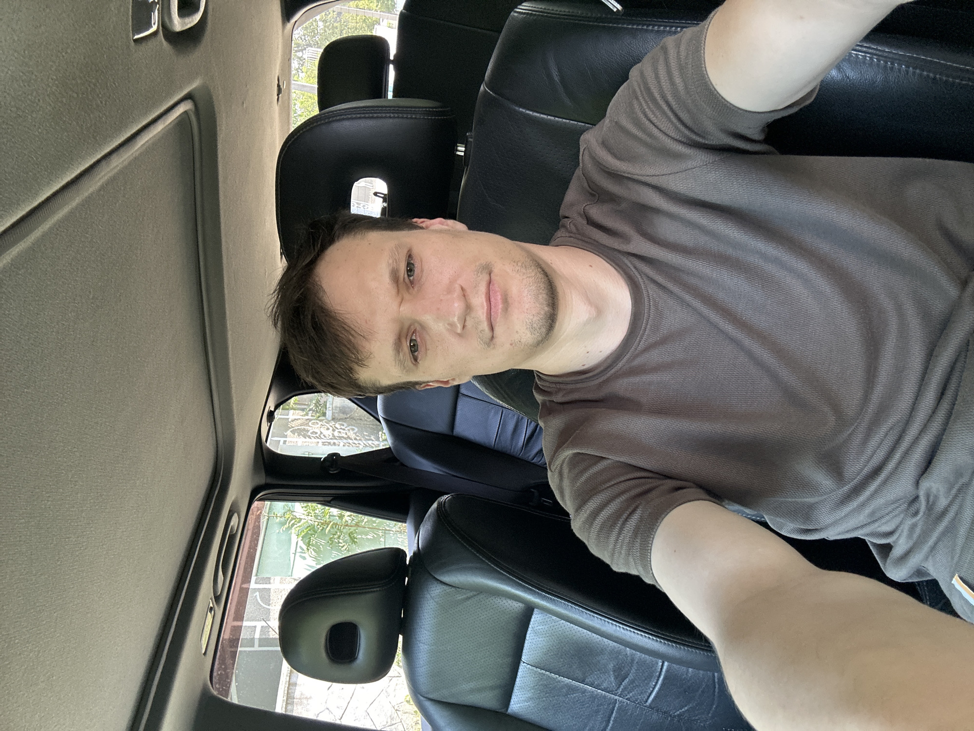 A man is sitting in the driver's seat of a car, in Makiv, Ukraine. He is wearing a grey t-shirt and has short brown hair. The car has black leather seats and a sunroof. The windows are open, and greenery and a stone wall are visible outside. The image is a selfie, taken from the man's perspective.