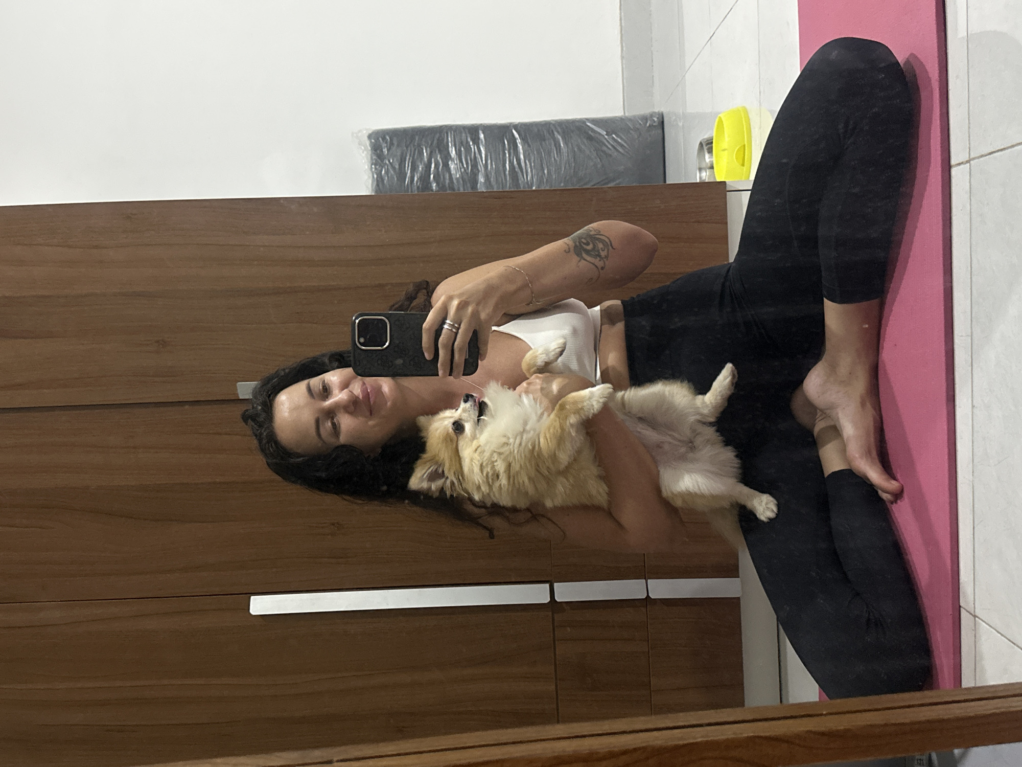 In Kuta Selatan, Indonesia, a woman with dark, wavy hair is seated on a pink yoga mat, holding a small, fluffy Pomeranian dog. She is wearing a white sports bra and black leggings. The woman is looking at her smartphone, which she is holding in her right hand, appearing to take a selfie. Her left arm is supporting the dog. The background shows a wooden wardrobe and a white wall. To the right of the woman, a rolled-up object wrapped in black plastic is visible. A yellow pet bowl is on the floor near the object. The lighting suggests it is daytime, and the scene appears to be indoors.