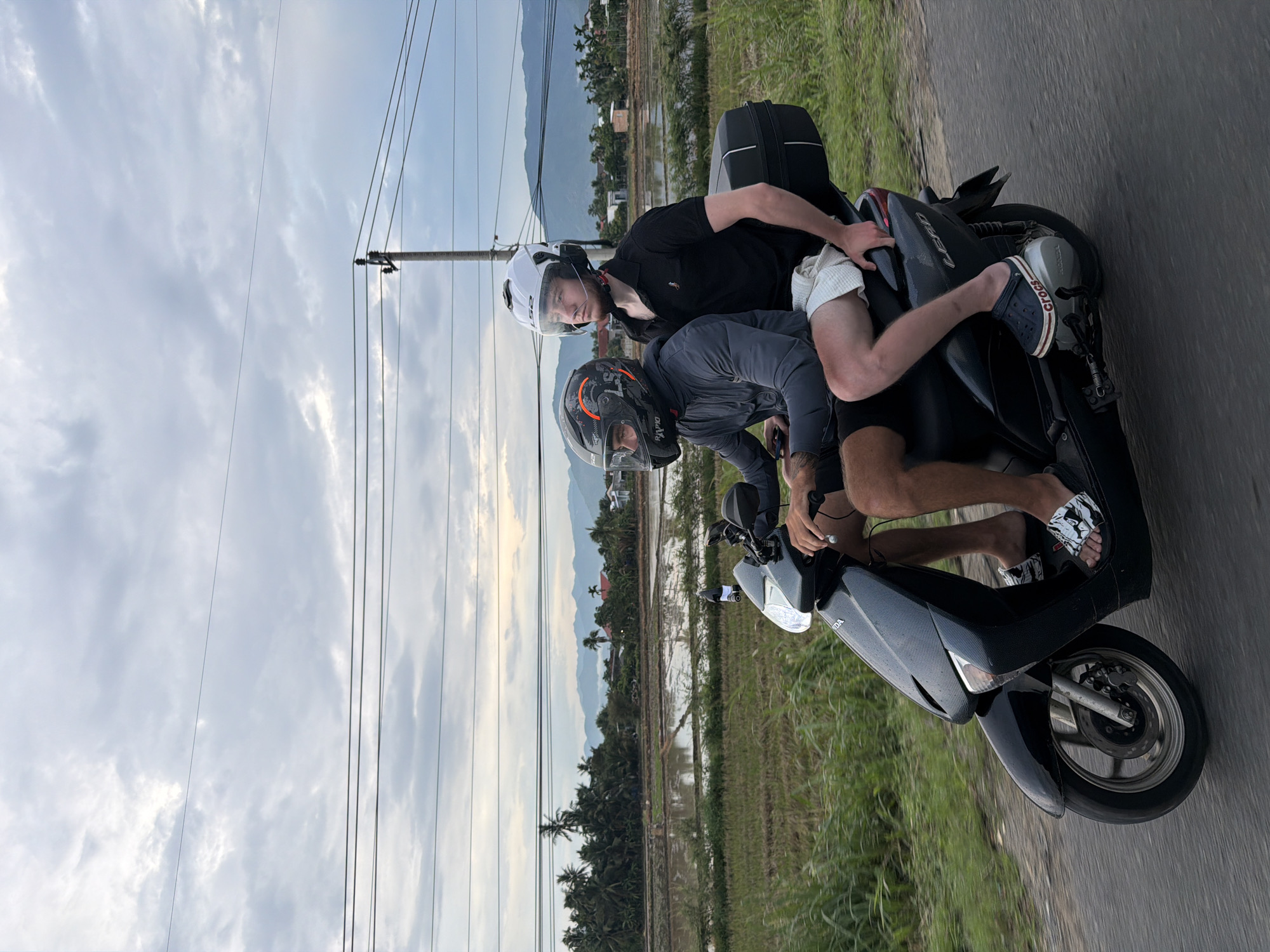 Two individuals are riding a black scooter on a paved road, traveling from left to right. The person in the front is wearing a gray hooded jacket and a dark helmet with orange accents. The person in the back is wearing a black polo shirt and white shorts, with a white helmet. Both individuals are wearing helmets. The scooter has a black storage box mounted on the rear. The background shows a rural landscape with rice paddies, palm trees, and distant mountains under a cloudy sky. Power lines are visible in the upper portion of the image. The scene is set in Dien Lac, Vietnam.