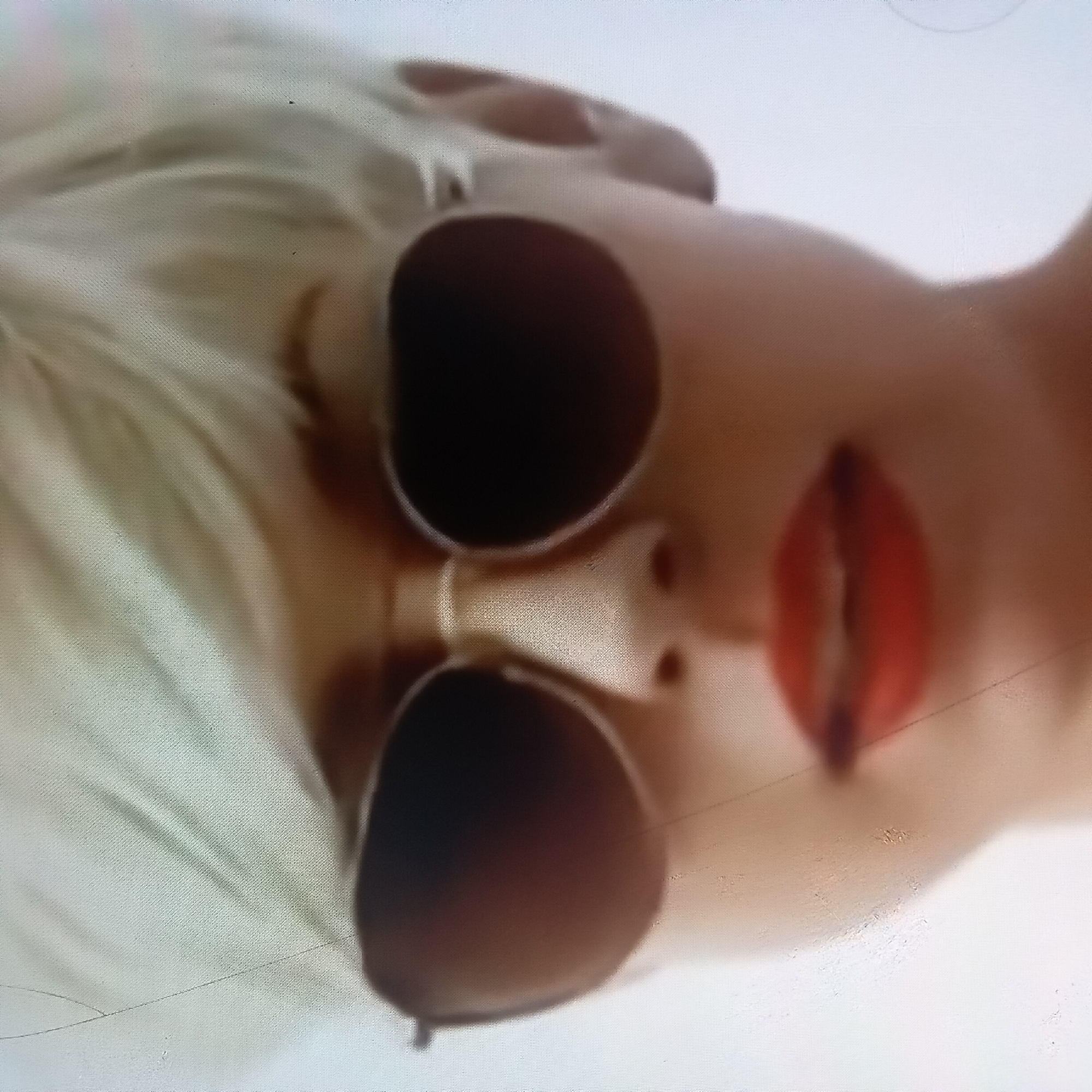 The image shows a close-up, slightly blurry, overhead view of a person's face wearing sunglasses. The person has blonde hair, and their lips are painted red. The image is taken at an angle, with the face tilted to the right. The background appears to be a neutral, light-colored surface, possibly fabric or a wall, with some subtle shading. There is no indication of a specific location in Almaty, Kazakhstan, or any other city. The overall impression is of a stylized portrait or fashion shot, with the focus on the face and accessories.