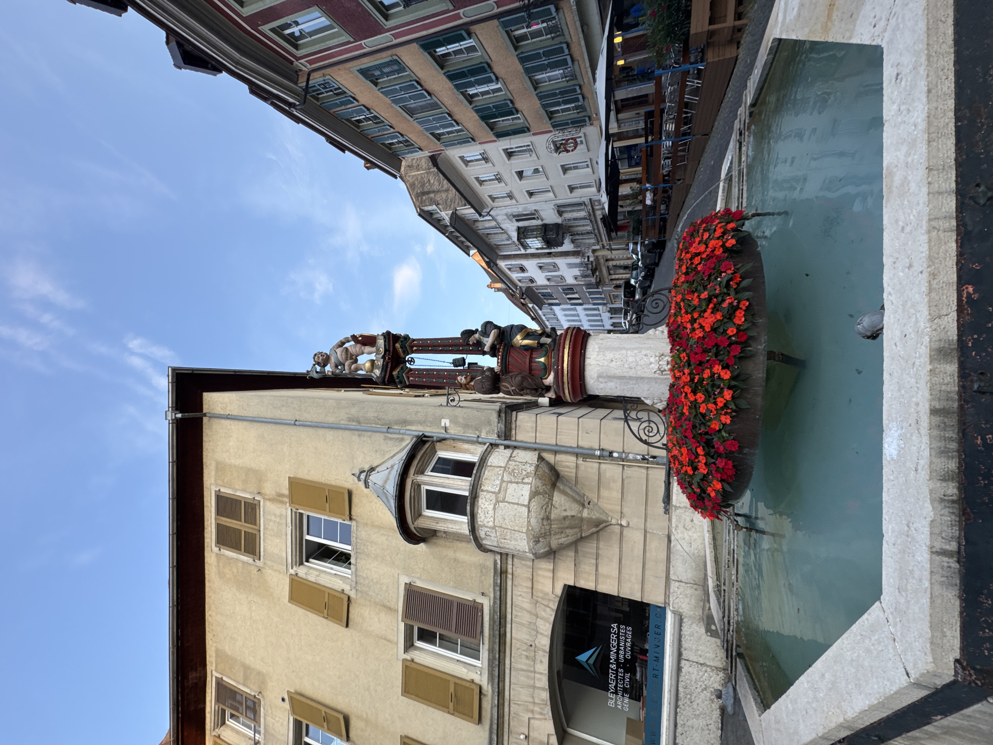 This image depicts the historic fountain located in the market square of Porrentruy, Switzerland. The fountain features a central sculpture with figures and is surrounded by a pool filled with clear, light blue water. Red flowers are densely arranged around the top of the fountain's basin, creating a vibrant contrast. The fountain is situated in front of an old, light-yellow building with several windows and brown shutters, some of which are open. To the right of the fountain, a street lined with colorful buildings, typical of Swiss architecture, recedes into the distance. These buildings have multiple stories, various colored facades, and numerous windows with shutters. An outdoor seating area with umbrellas and tables is visible along the sidewalk in front of some of the buildings, suggesting a cafe or restaurant. The sky above is blue with scattered white clouds. The scene is a street-level, medium-angle shot, capturing the fountain and its urban surroundings.