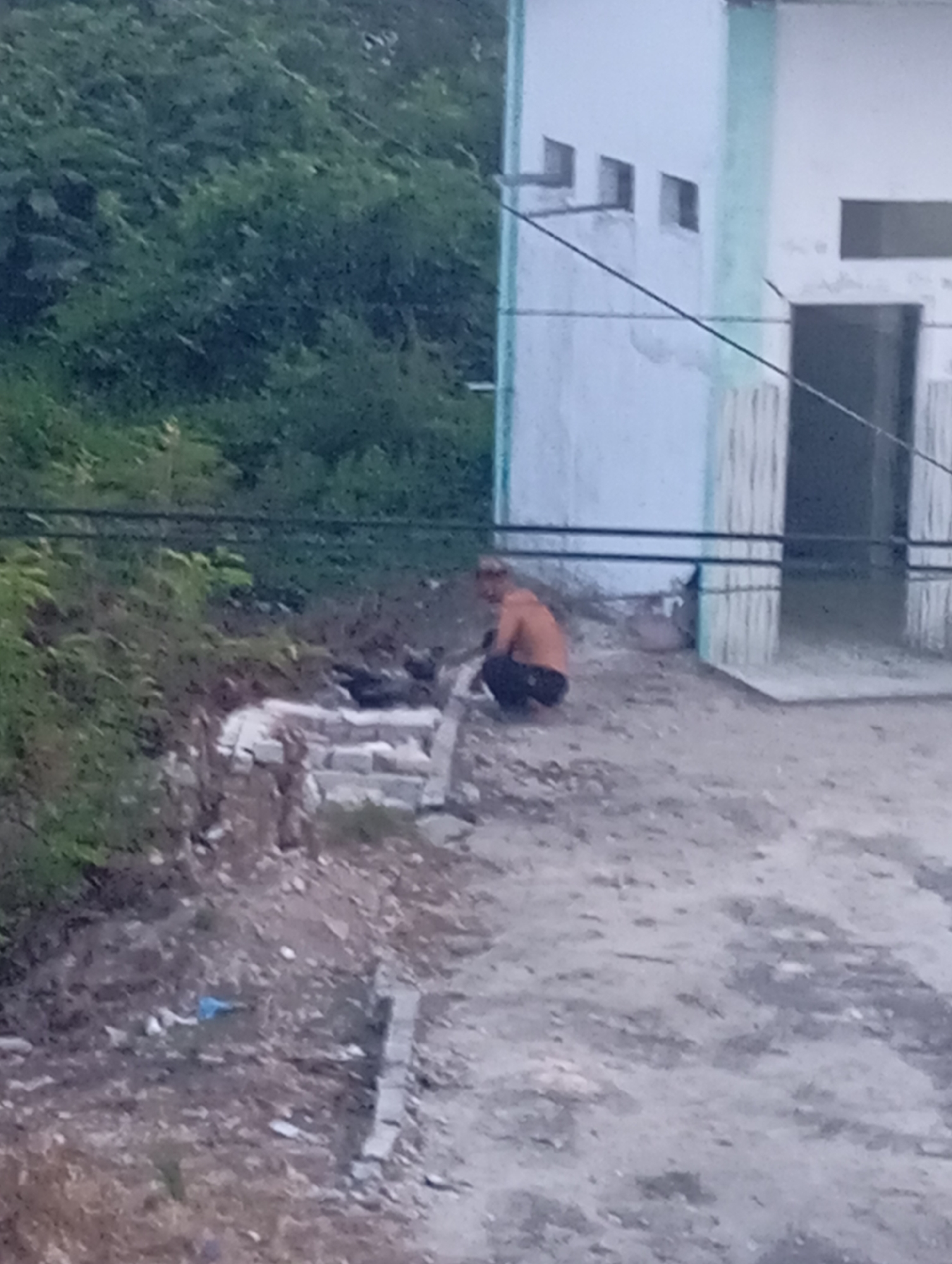 The image depicts an outdoor scene with a man squatting on the ground near a partially constructed building. The man, appearing to be shirtless and wearing dark pants, is positioned to the left of the frame, amidst scattered debris and what looks like a pile of bricks. Behind him, a white building with light green trim is visible, featuring several small rectangular openings or windows near the roofline. The ground is a mix of dirt and gravel. Lush green foliage is present in the background, suggesting a vegetated area adjacent to the construction. The overall impression is one of a work-in-progress or an undeveloped area.