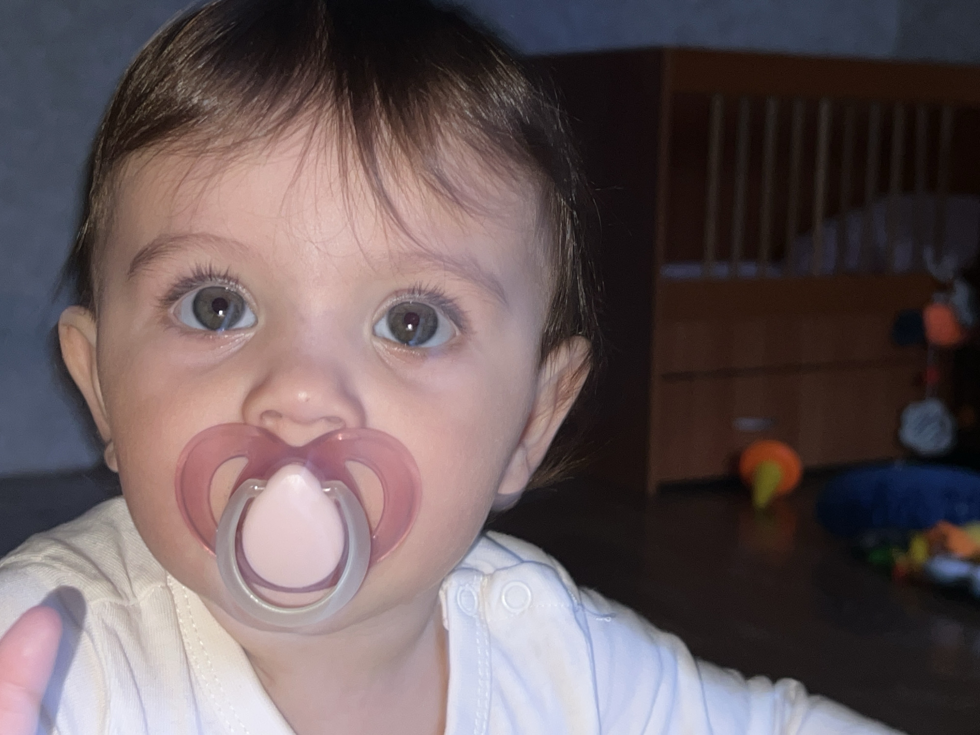 An infant is depicted in a medium close-up, situated indoors within what appears to be a domestic residence in Elena, Bulgaria. The infant, with dark brown hair and gray-green eyes, is wearing a light-colored, long-sleeved garment with shoulder snaps. A pink and clear silicone pacifier is actively in the infant's mouth. The infant's gaze is directed forward, toward the camera. In the background, to the right of the infant, a dark wooden crib with vertical slats is partially visible. The floor in the background displays several blurred objects, including a distinct yellow and orange conical toy and a blue mat. Lighting is subdued, suggesting an indoor evening or low-light condition.