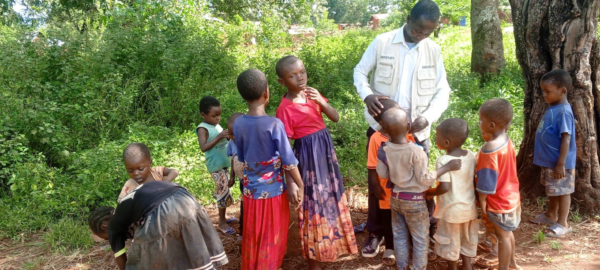 The image shows a group of children and a man in an outdoor setting in Ngalula, Democratic Republic of the Congo. The man, wearing a light-colored vest with text, appears to be interacting with a cluster of children, some of whom are looking at him. Other children are situated around them. The children are wearing an assortment of clothes, including dresses, shirts, and pants. The environment includes trees, greenery, and a dirt ground.