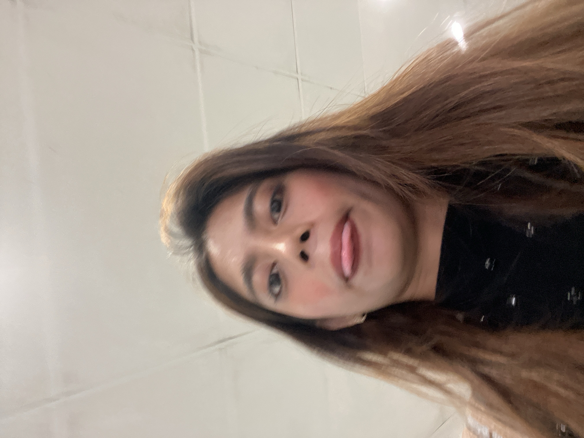 A close-up, low-angle selfie features a young woman looking upwards towards the camera, her head tilted diagonally across the frame. She has long, straight brown hair that flows to the right. Her expression is playful, with her tongue slightly sticking out, and she wears makeup including blush on her cheeks. Her eyes are dark and focused on the lens. She is dressed in a dark-colored top, possibly black, visible on the right side of the image, featuring subtle shiny embellishments. The background is a plain, light-colored indoor surface, likely a wall or ceiling, with faint intersecting lines suggesting panels or tiles. The image conveys a lighthearted and informal moment, set indoors in Carmen, Philippines.