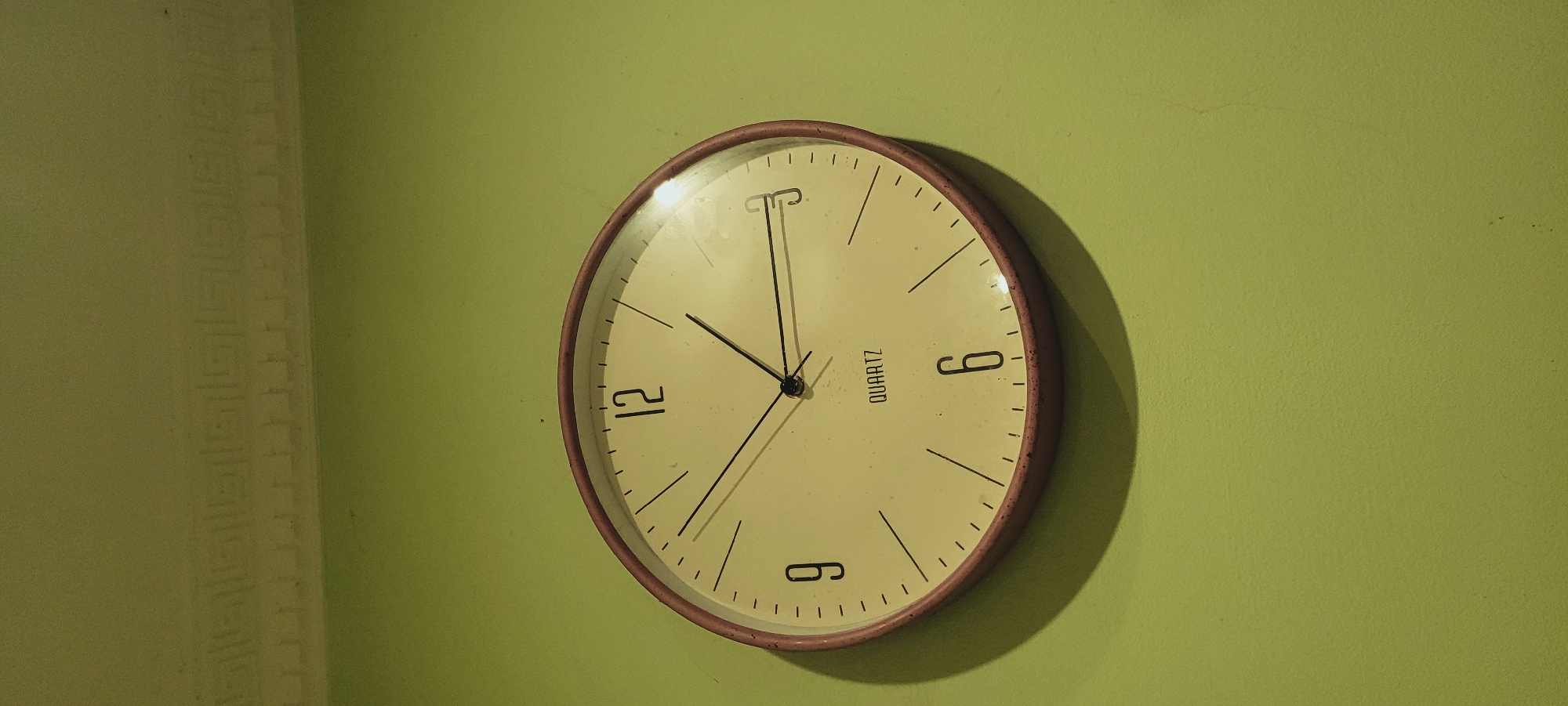 A round, analog wall clock with a cream-colored face and a thin, reddish-brown bezel is mounted on a light green wall. The clock has black hour markers and Arabic numerals at 12, 9, and 6. The hour hand points just before the 10, the minute hand points at the 1, and the second hand is slightly past the 12. The word "QUARTZ" is printed in the center of the clock face. There is a decorative molding along the top edge of the wall. The lighting casts a shadow of the clock on the wall to its left.