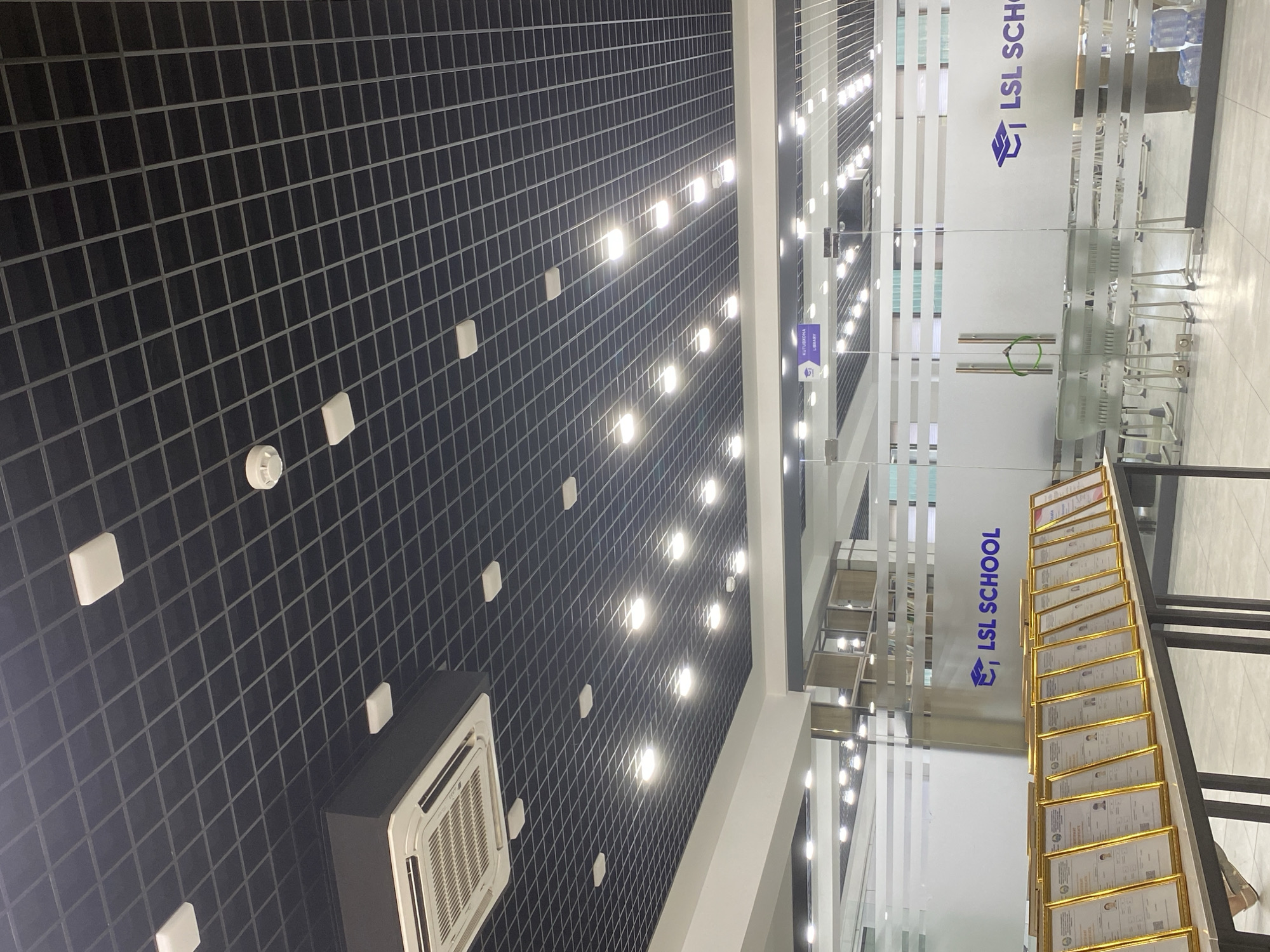 The image shows the interior of a modern building, likely a school or educational institution, with a grid-patterned dark ceiling containing recessed lighting and a visible air conditioning unit. Below the ceiling, a glass-partitioned entrance leads to an area with chairs and tables. The glass doors and walls are frosted with the text "LSL SCHOOL" and a graduation cap logo in purple. In the foreground, a table displays a row of framed certificates with gold frames, suggesting an award ceremony or recognition event. The overall impression is one of a clean, well-lit, and organized space. The location is identified as Namangan City, Uzbekistan.